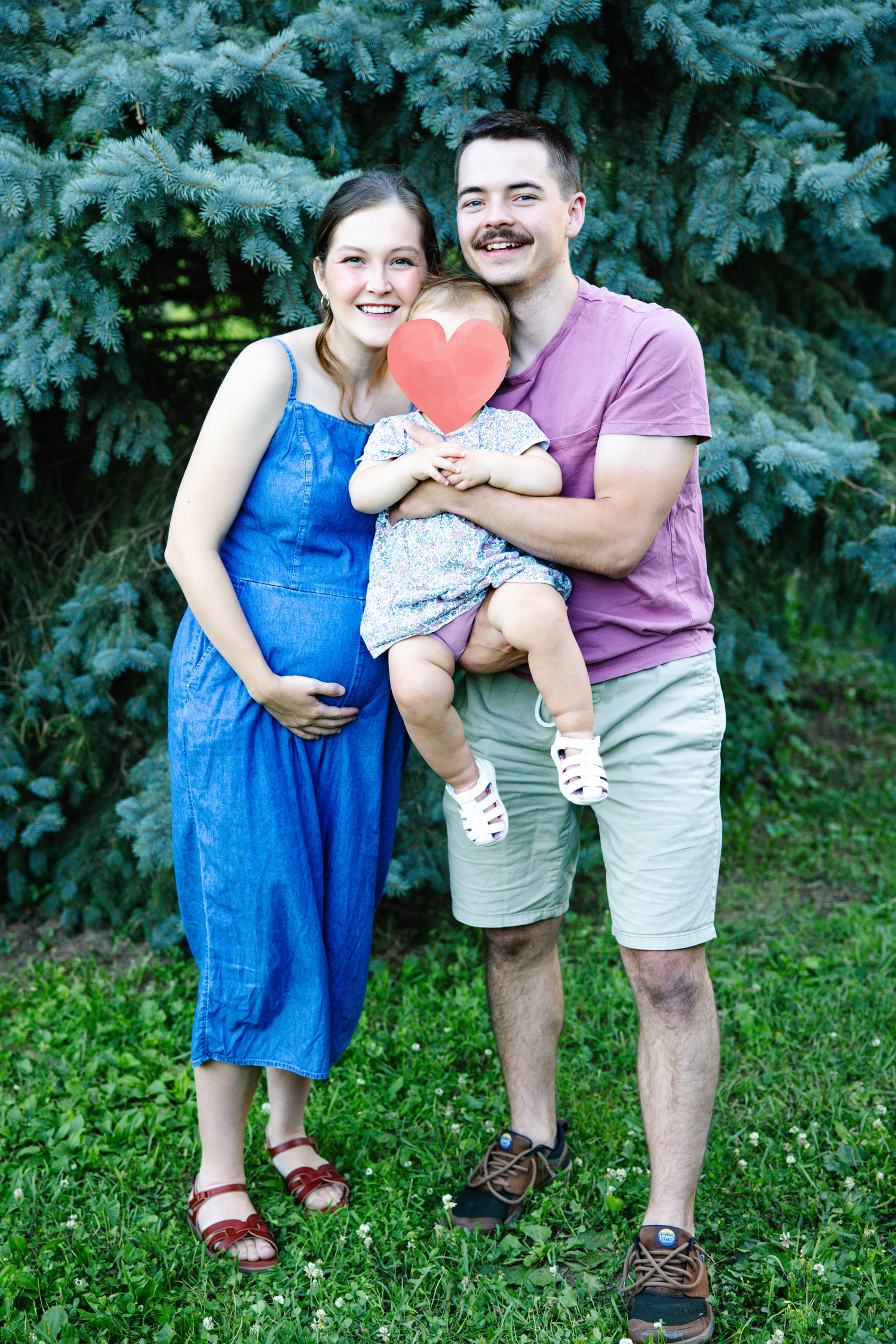 A pregnant woman in blue dress and man holding toddler with heart cutout pose outdoors near evergreen trees.