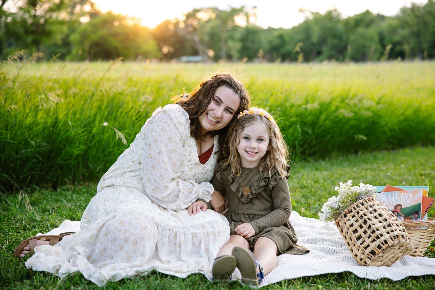 A mother and young daughter smile together on a white blanket in a sunny green field during a golden hour picnic.