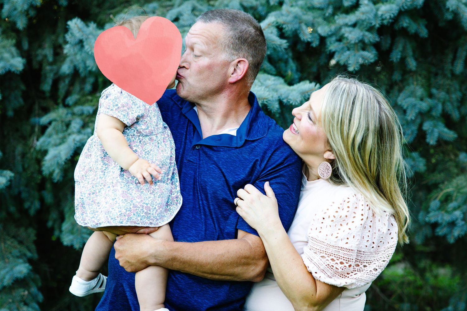 A couple shares a kiss over their baby girl holding a red heart, posing outdoors with green trees behind them.