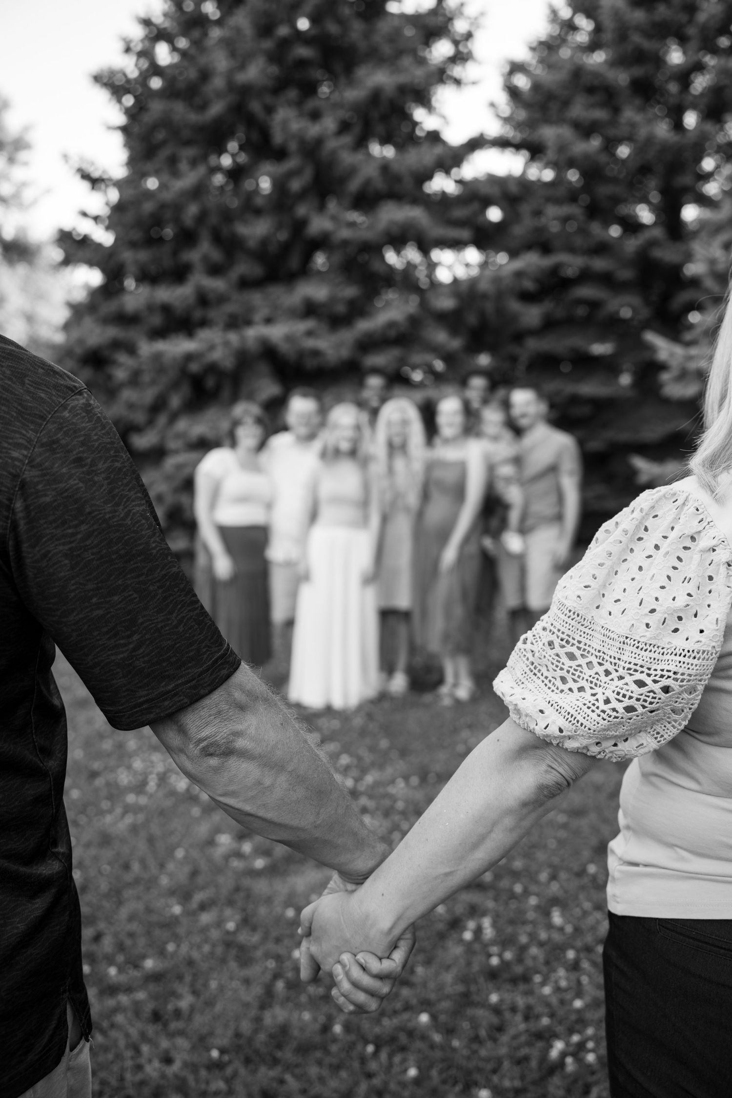 Black and white photo of couple holding hands facing a group of people gathered outdoors near tall evergreen trees.