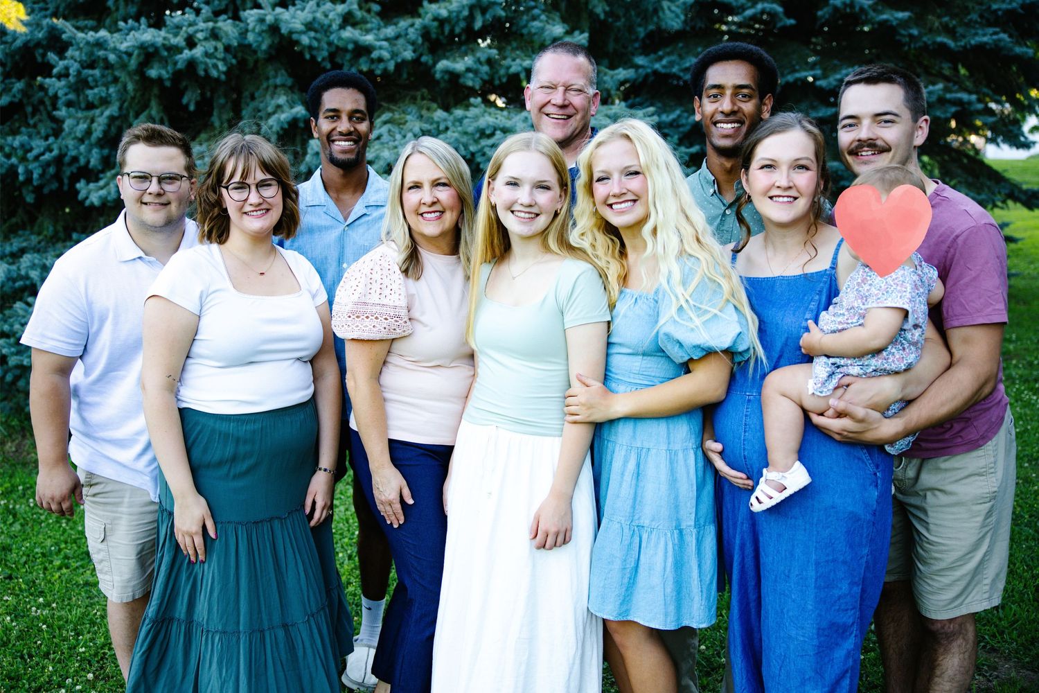 Large family group smiling outdoors in summer, posing together on green lawn with trees in background.