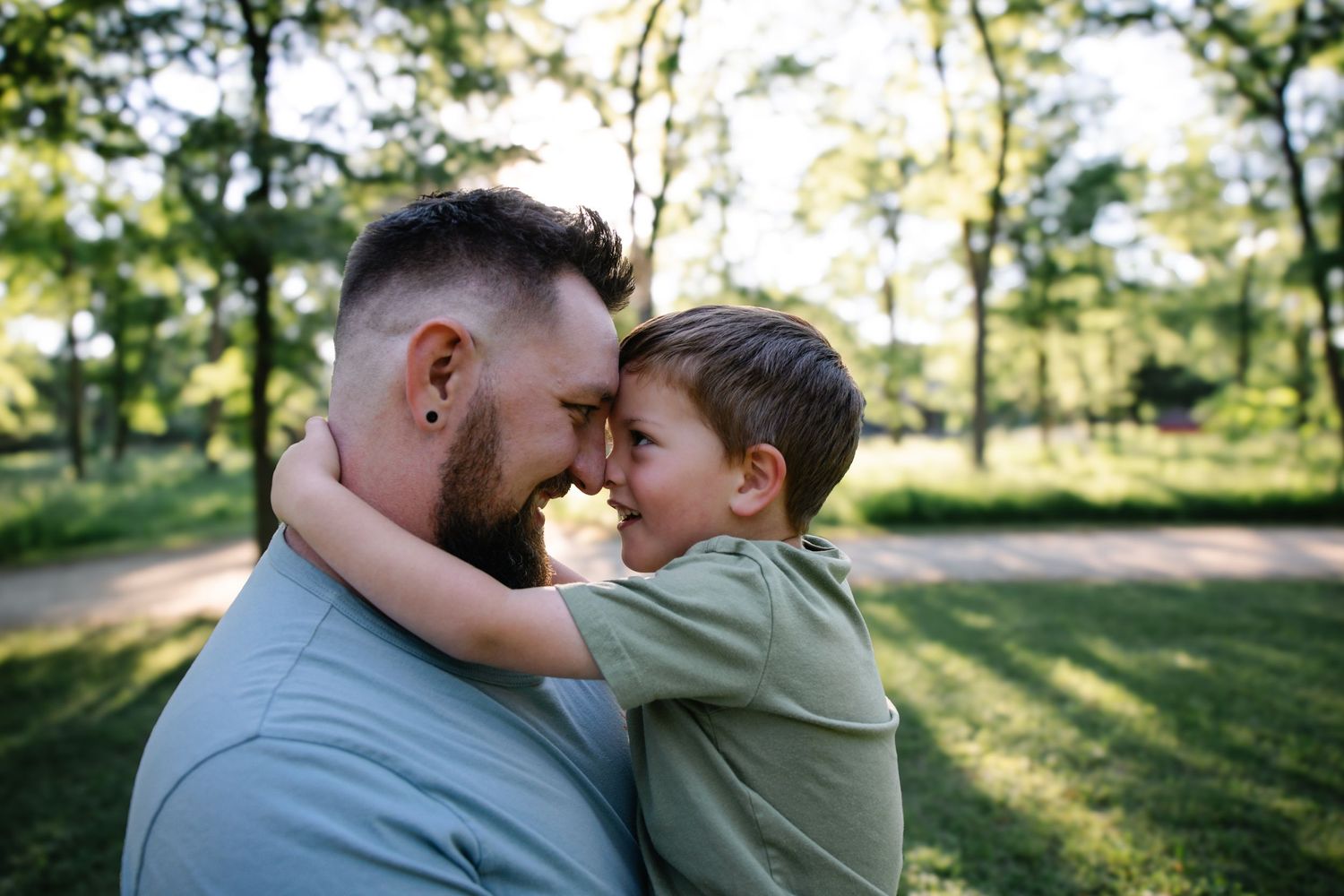 A loving father and young son touch foreheads in a tender embrace outdoors in a sunny green park.