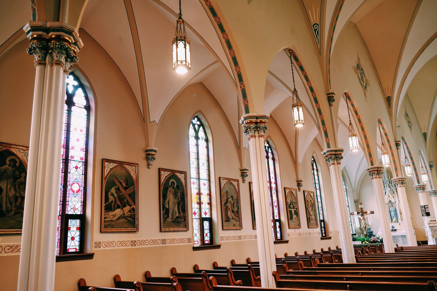 Gothic church interior with arched ceilings, stained glass windows, wooden pews, ornate columns, and religious paintings lining the walls.