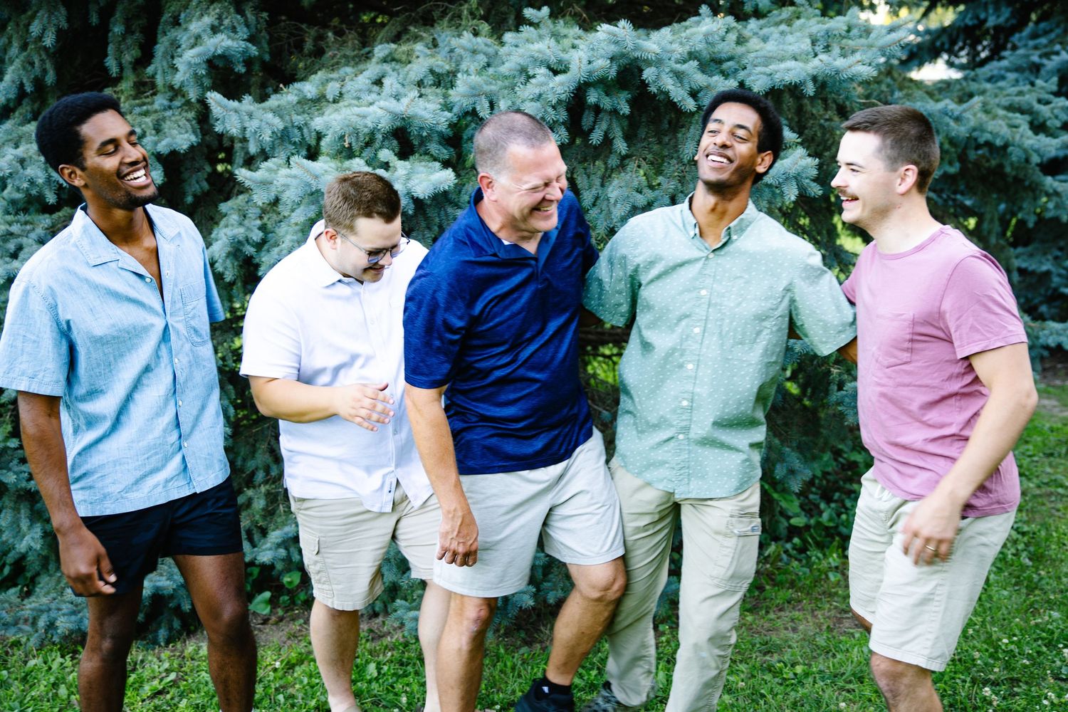 Five men laughing and smiling together outdoors in front of evergreen trees, wearing casual summer clothing.