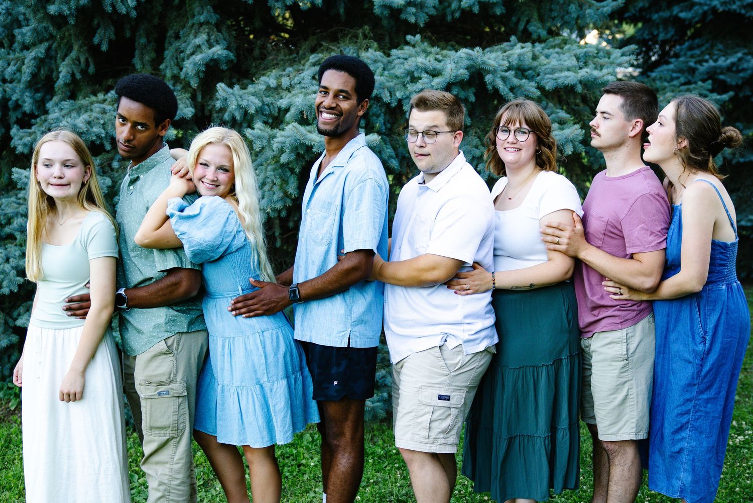Group of eight young adults posing together outdoors in front of evergreen trees, dressed in casual summer attire.