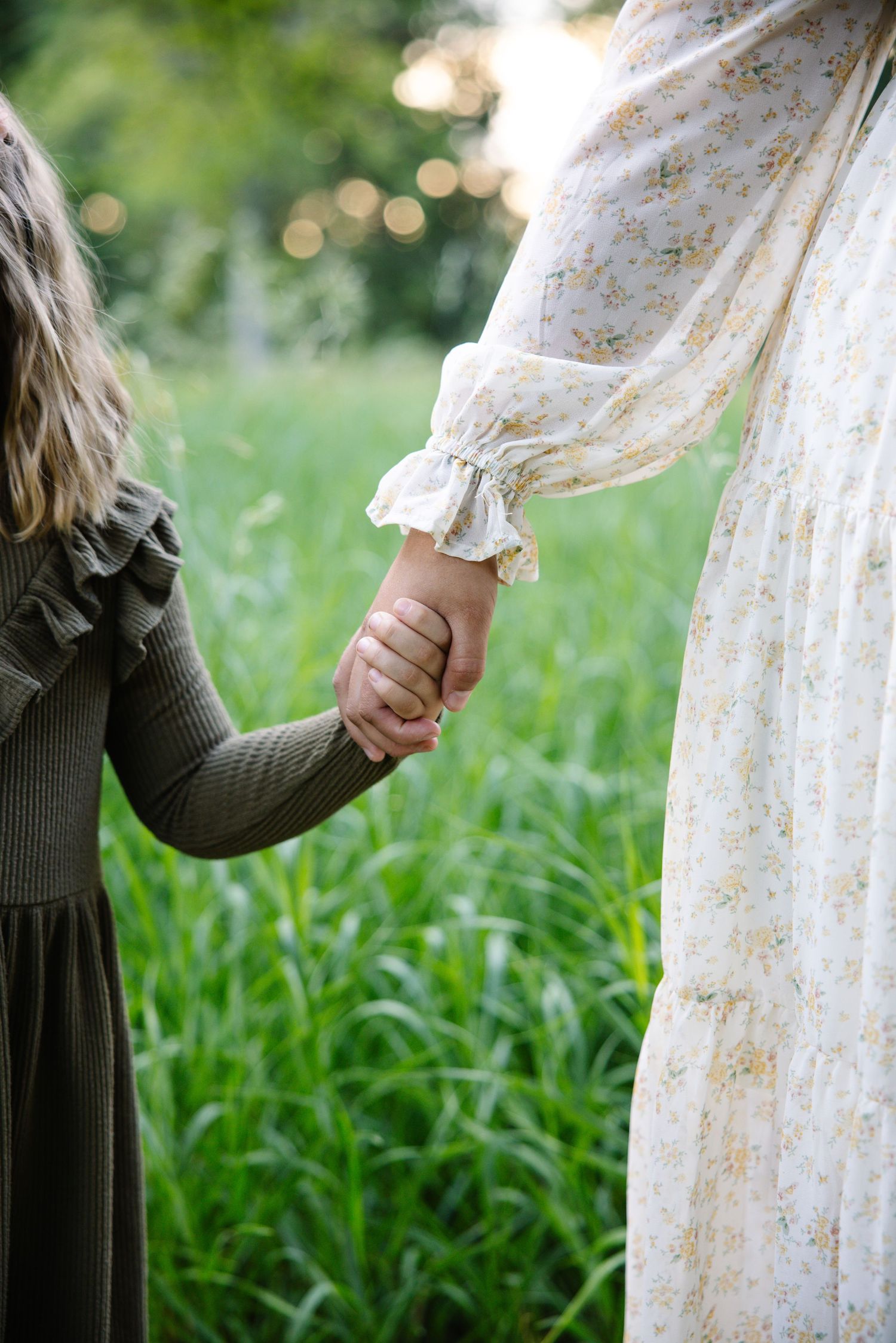 A mother and young girl hold hands in a lush green field, wearing floral and olive dresses.
