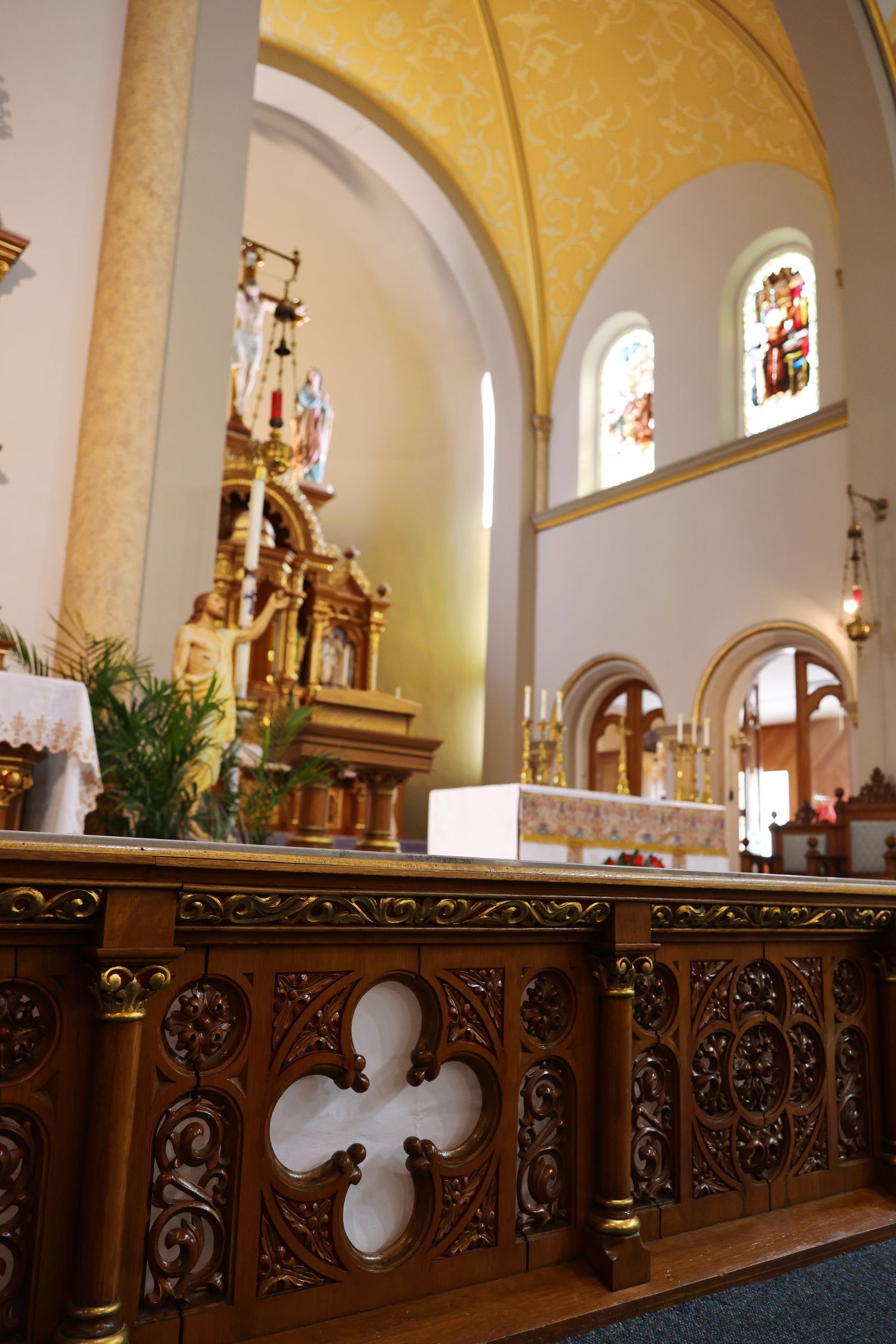Ornate wooden church altar rail with carved quatrefoil details, golden altar, stained glass windows, and arched ceilings.