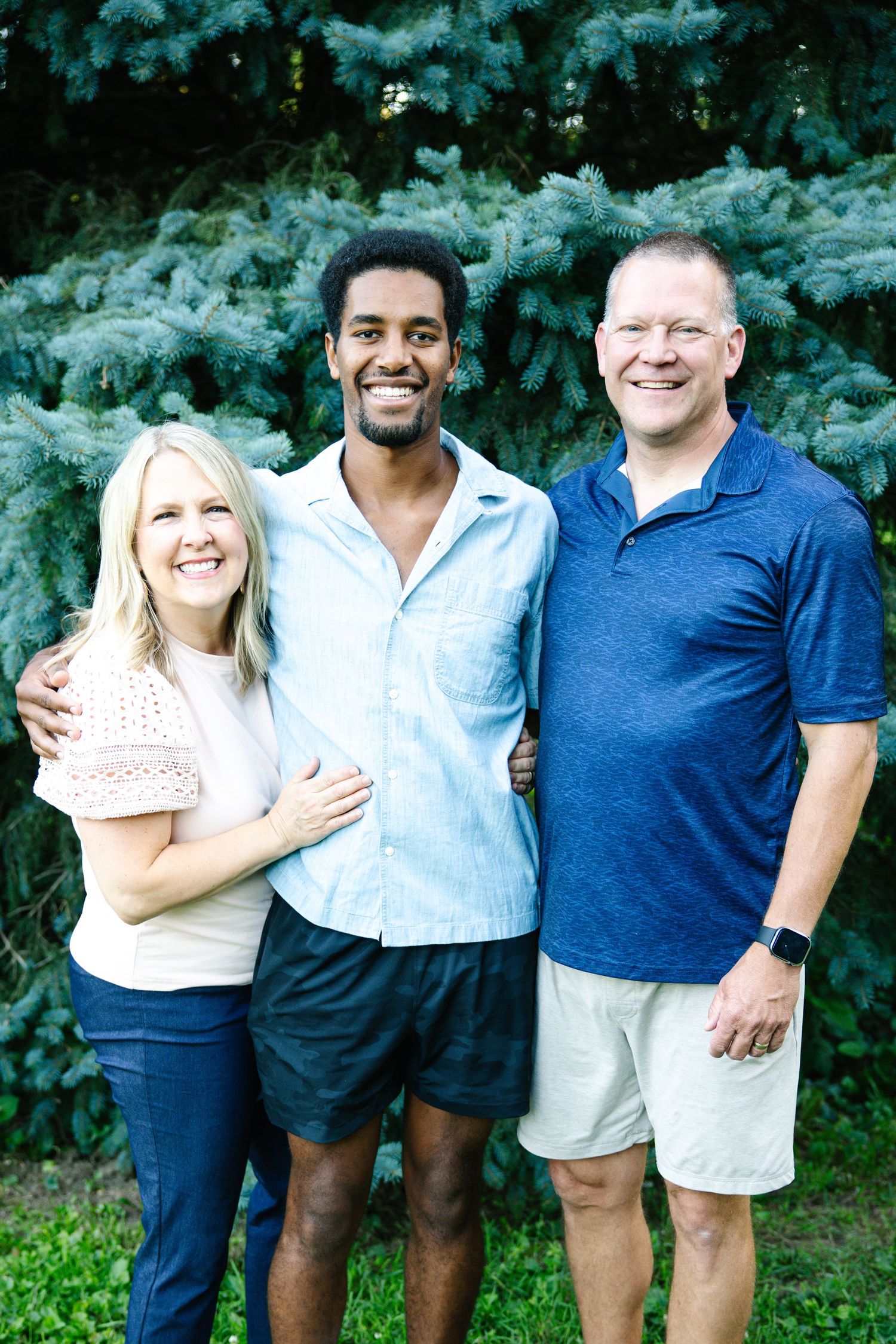 Three smiling people posing outdoors in front of evergreen trees on a sunny day.