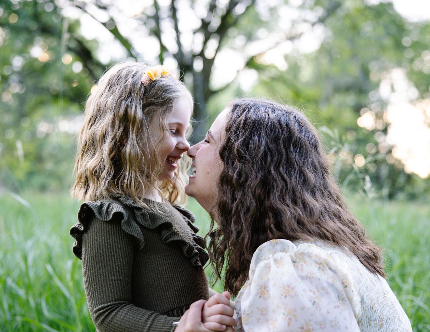 A mother and young daughter share a tender nose-to-nose moment outdoors in a lush green field.