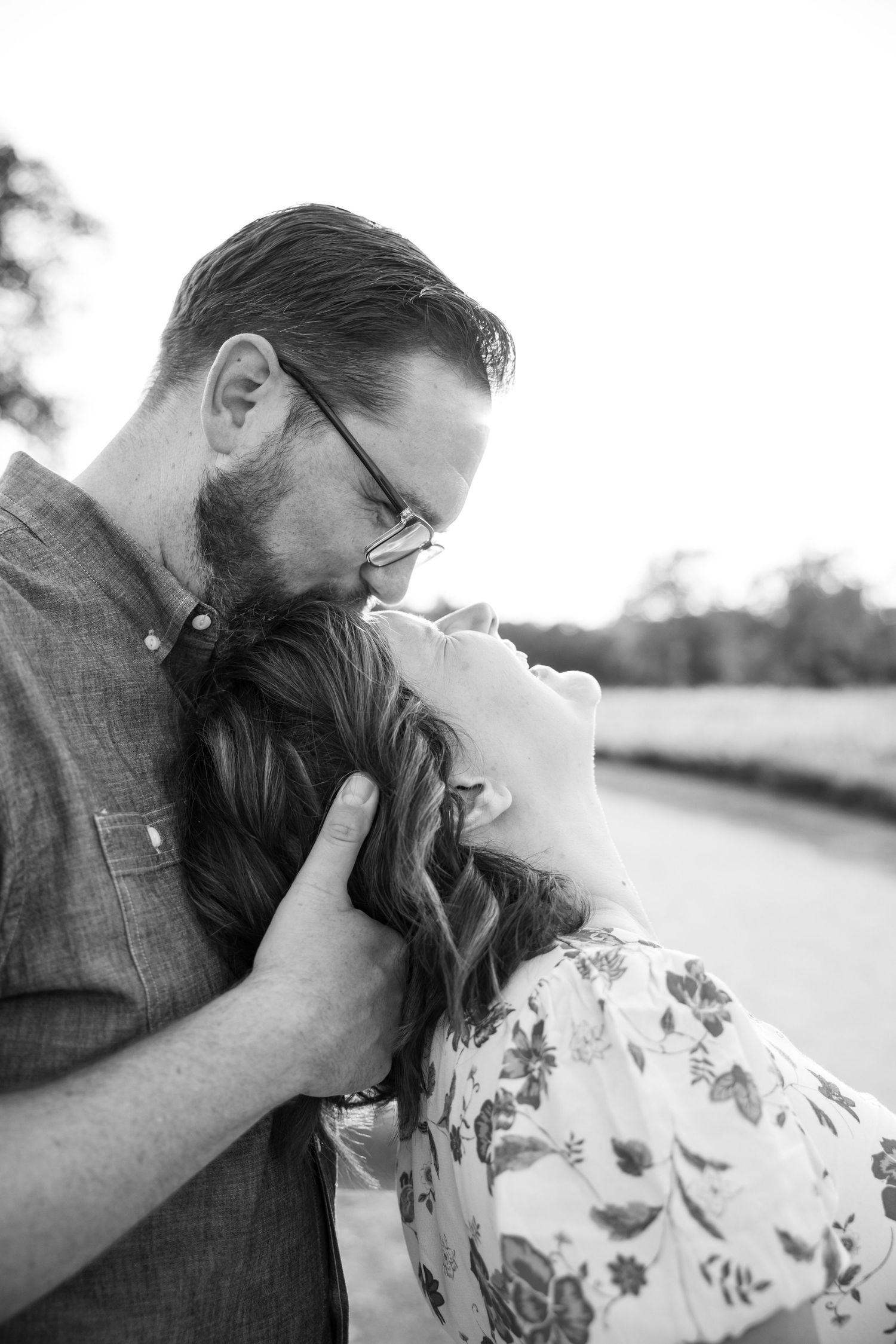 Black and white photo of a bearded man with glasses leaning in to kiss a woman with long wavy hair outdoors.
