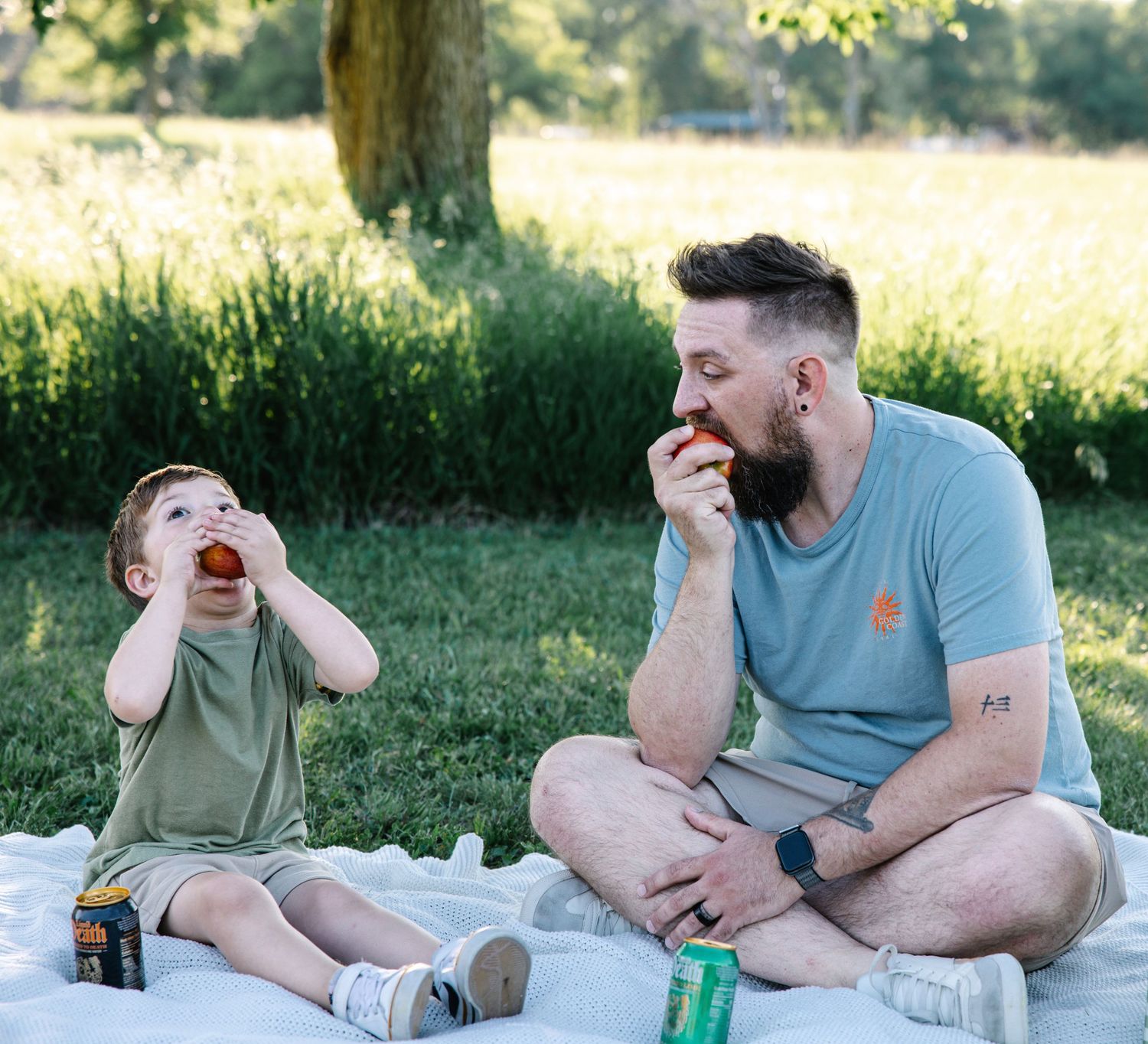 Father and young son sitting on blanket in park eating apples together on a sunny day.