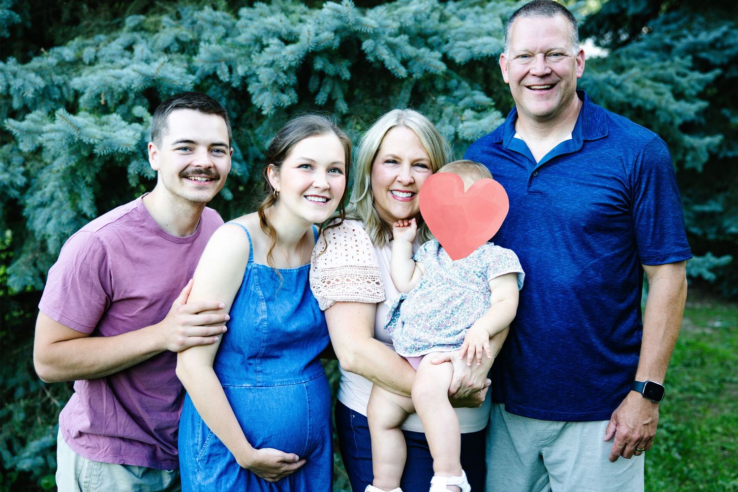 Happy family of four posing outdoors near evergreen trees, with a baby holding a red heart cutout.