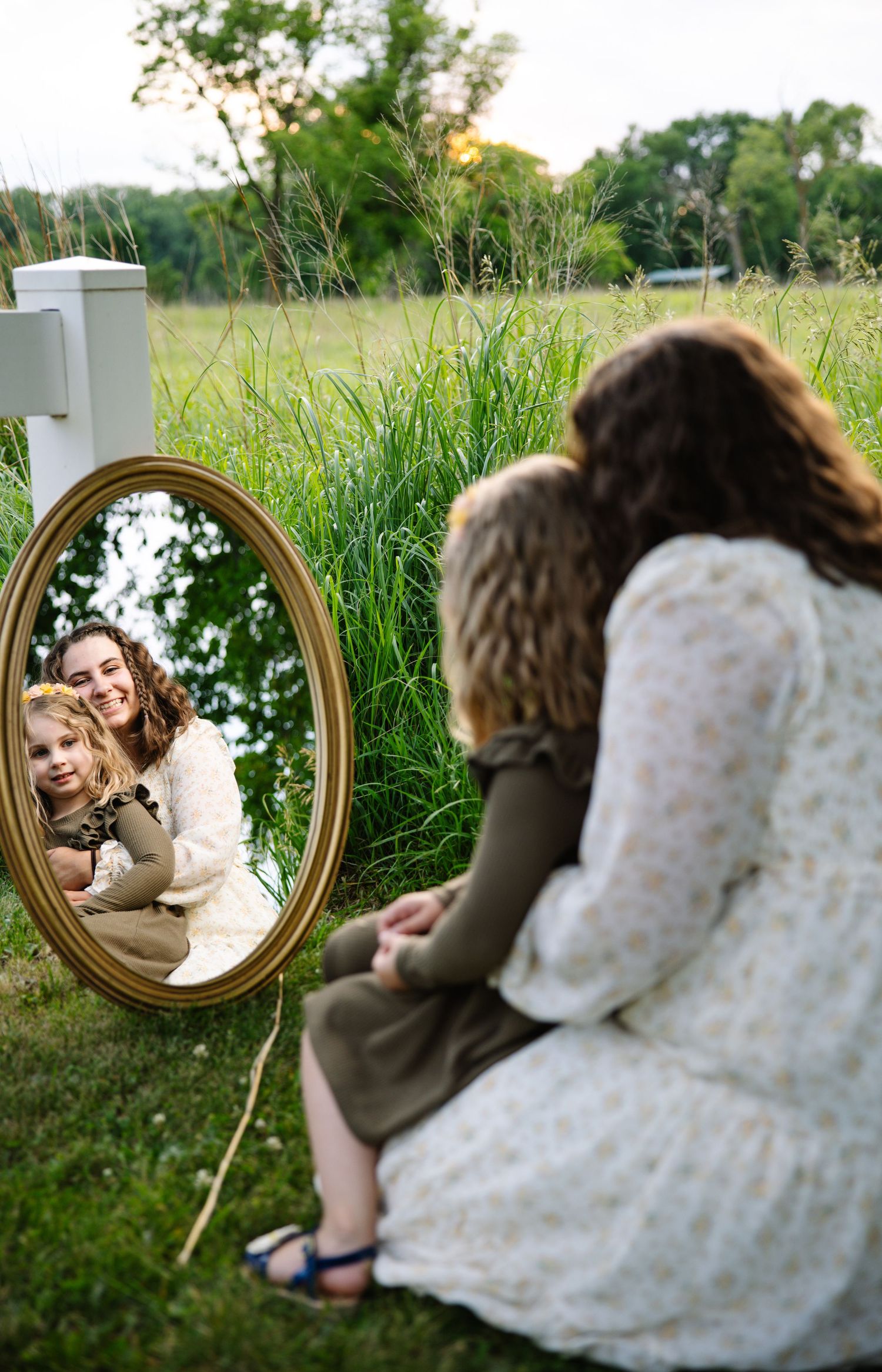A mother and child are reflected smiling in a gold oval mirror placed in a lush outdoor garden setting.