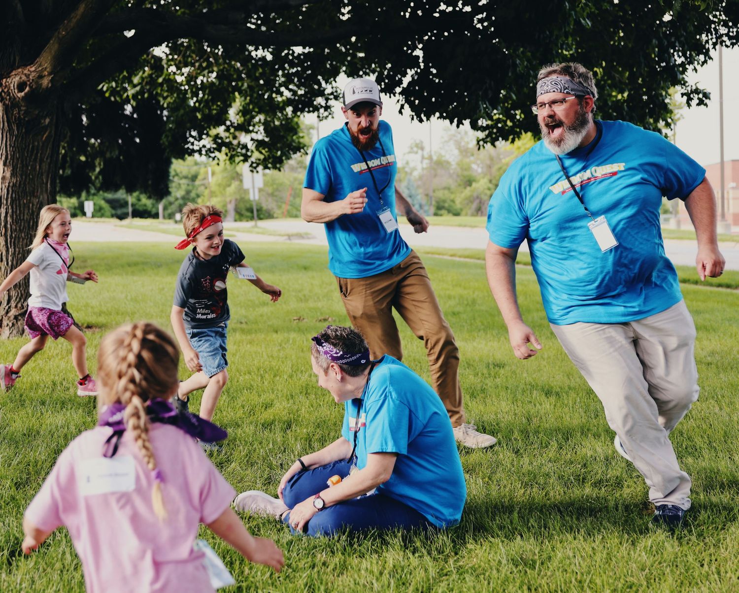 Adults and children playing outdoors on green grass near a tree during a fun group activity event.