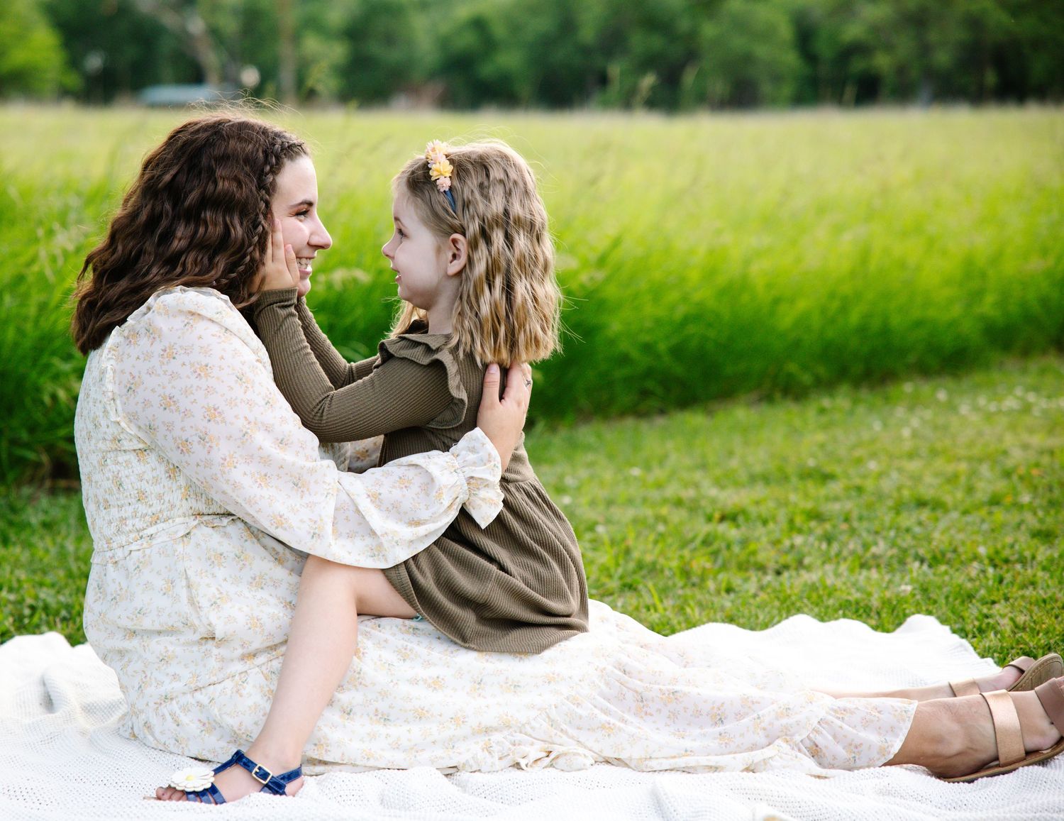 A mother and young daughter in matching floral outfits sit face-to-face on a white blanket in a green field.