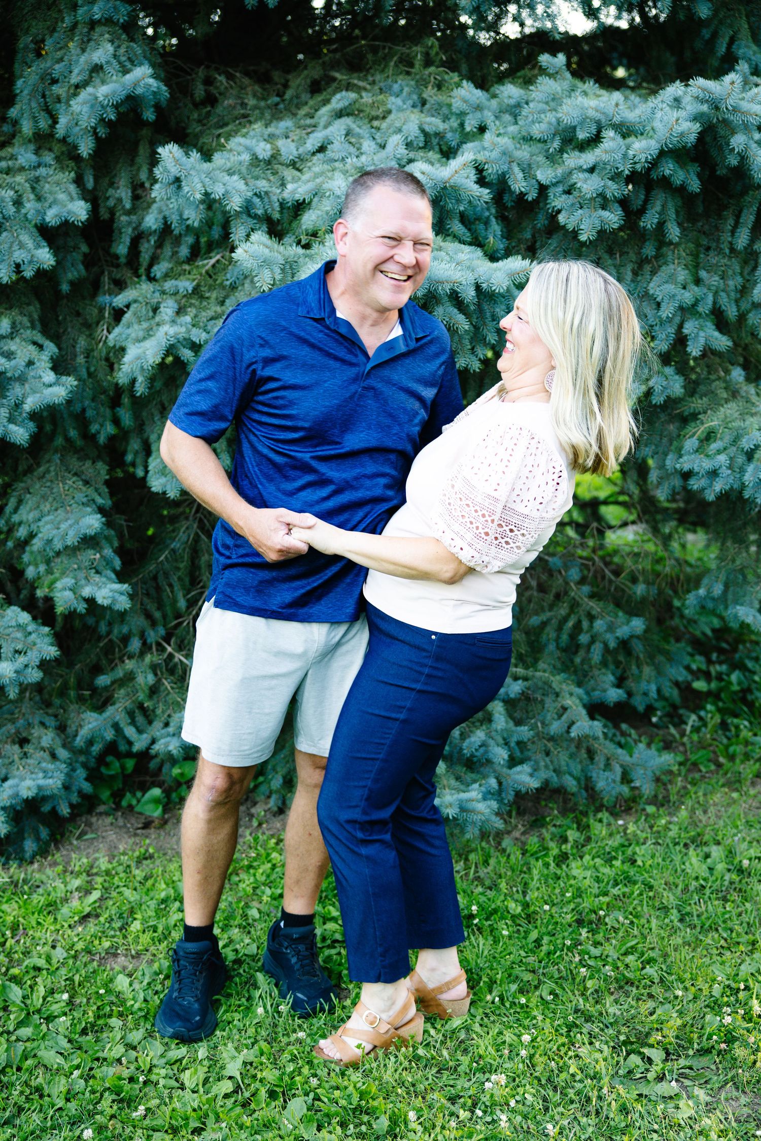 A happy couple holds hands and laughs together outdoors in front of evergreen trees in summer.