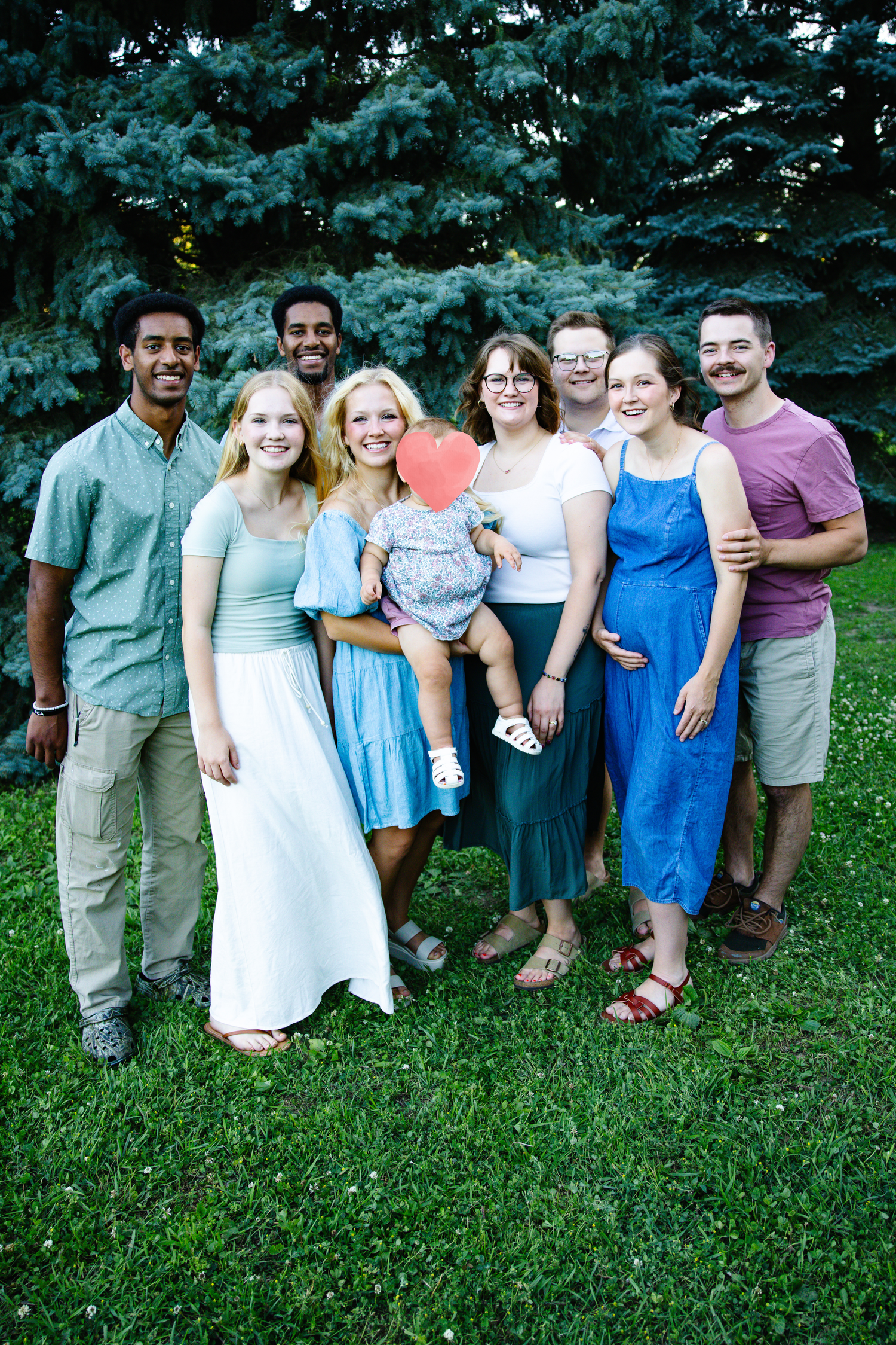 A diverse group of eight smiling adults and a child pose together outdoors on a sunny day near evergreen trees.