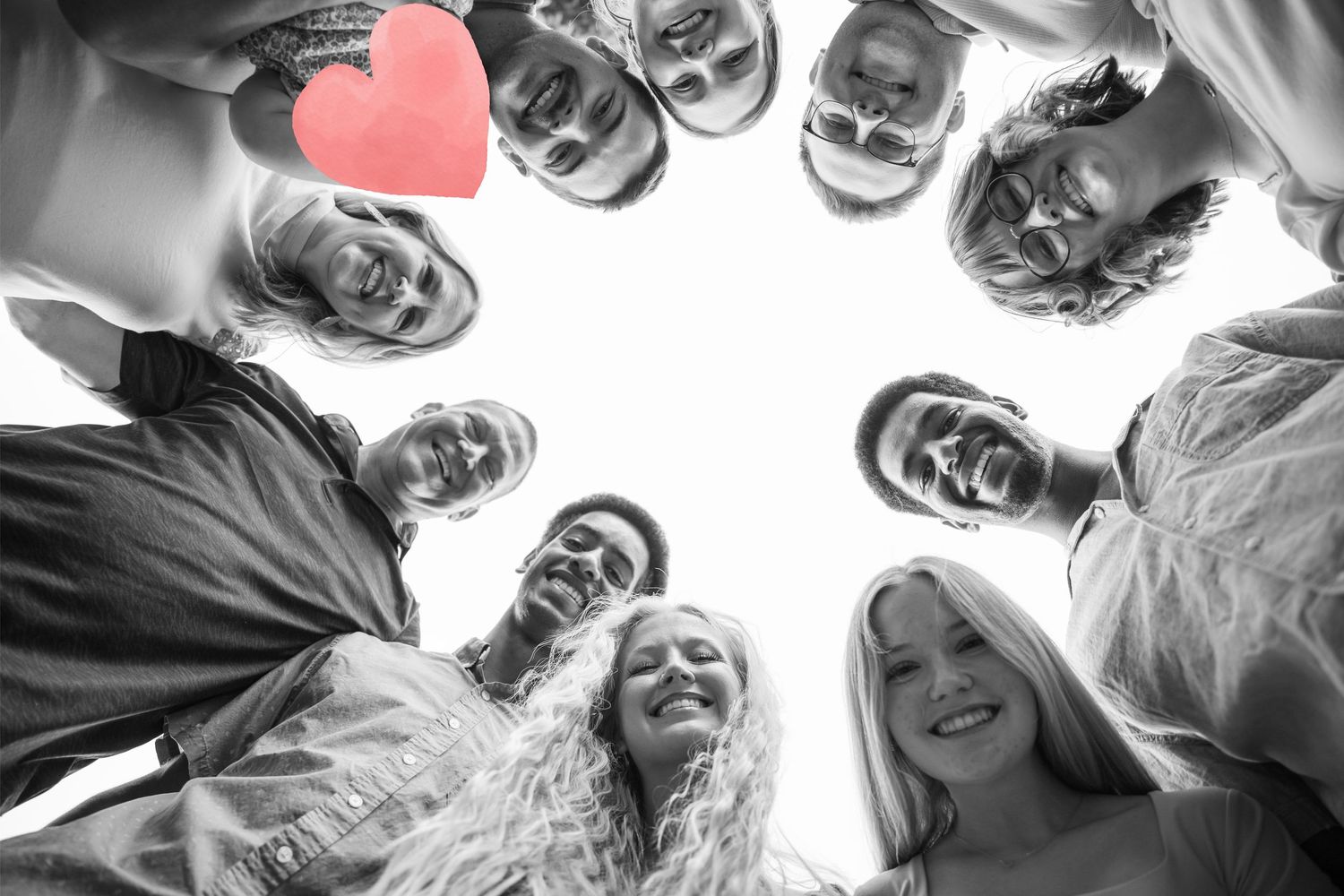 Black and white photo of a diverse group of smiling friends huddled in a circle, viewed from below, with a red heart.