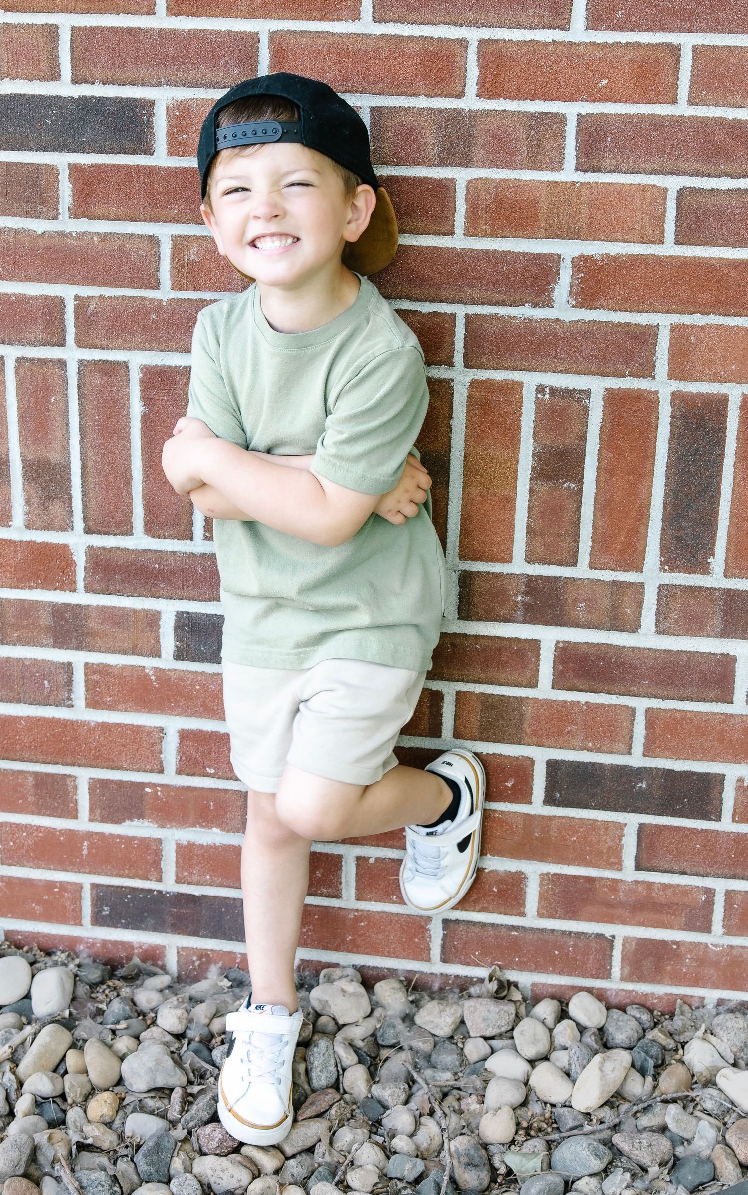Smiling young boy in green shirt, khaki shorts, and snapback cap leaning against a brick wall with arms crossed.