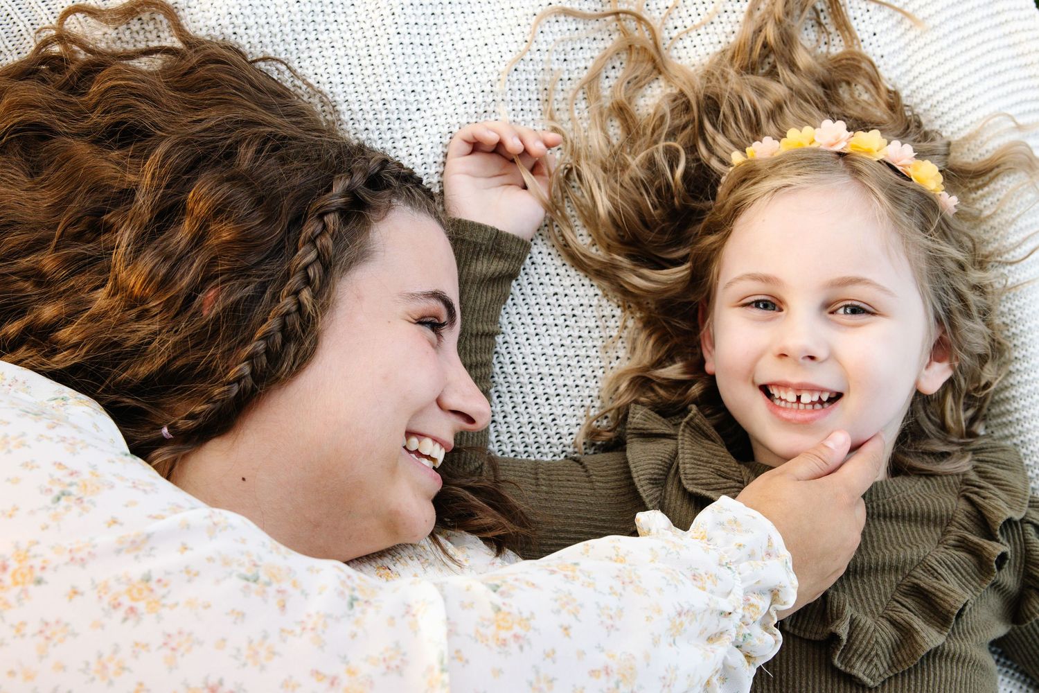 Two girls lying down laughing together, one with braids and one with a yellow flower headband.
