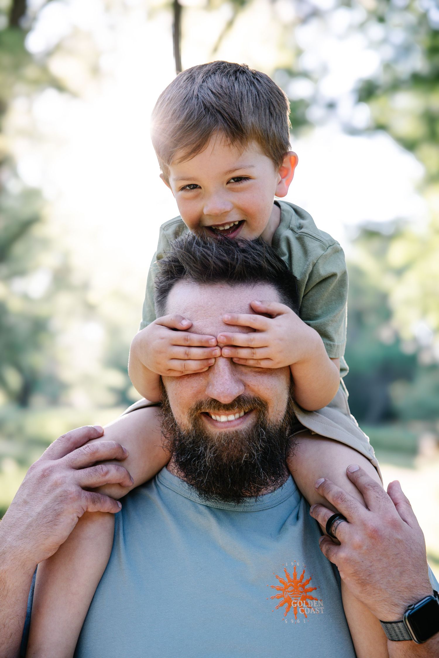 A laughing boy sits on his bearded father's shoulders covering his eyes in a sunny park.