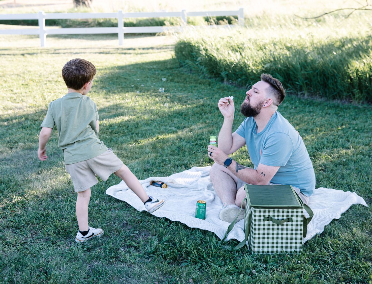 A bearded dad blows bubbles for his young son during a picnic on a grassy lawn.