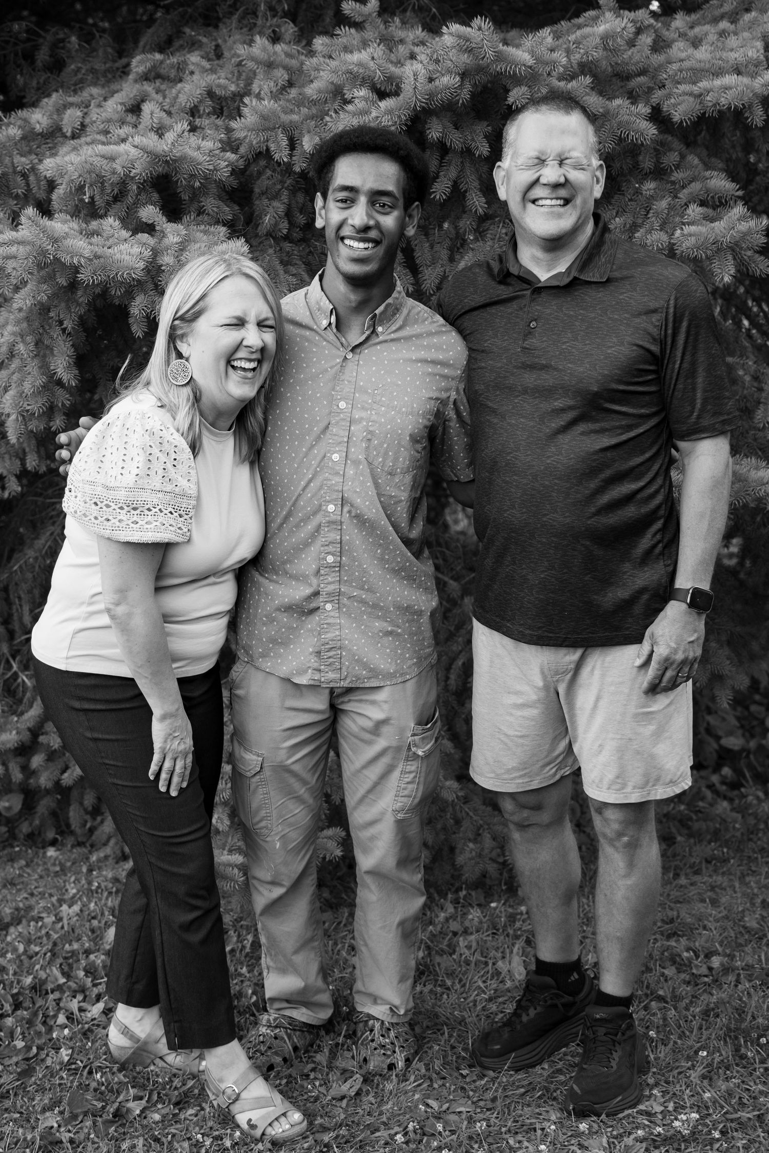 Black and white portrait of two adults and a young man laughing together outdoors near evergreen shrubs.