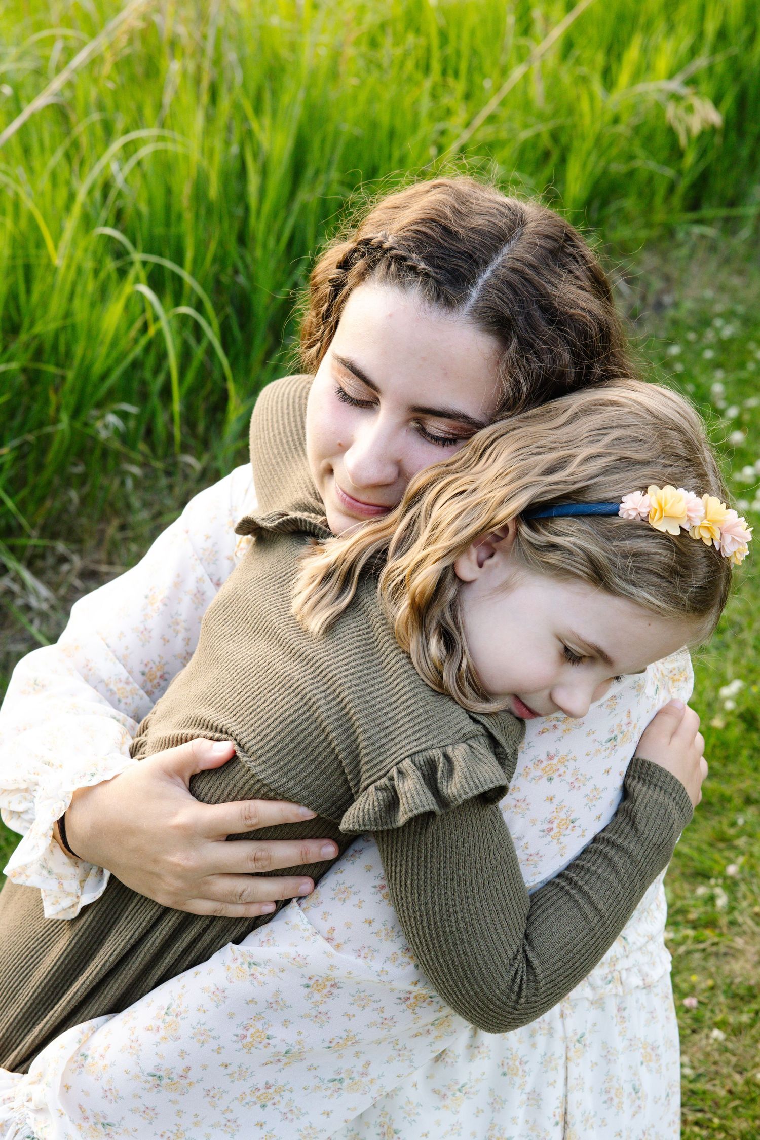 A tender moment as a mother gently hugs her young daughter in a sunlit green field.