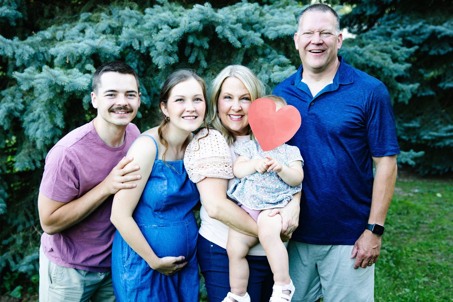 Happy family of four posing outdoors near evergreen trees, with a woman holding a baby whose face is covered by a red heart.