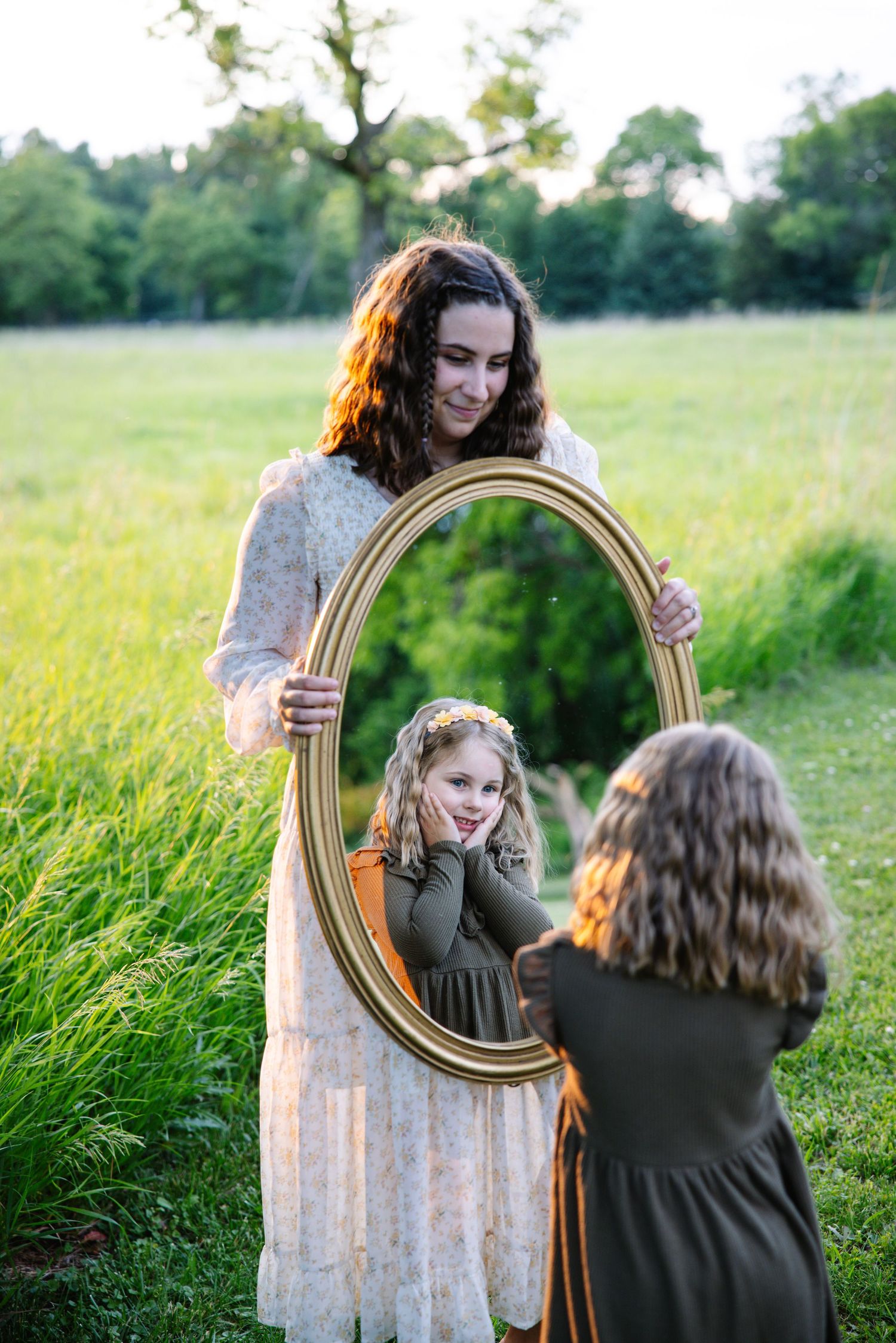 A woman holds a gold oval mirror reflecting a young girl admiring herself in a sunny field.