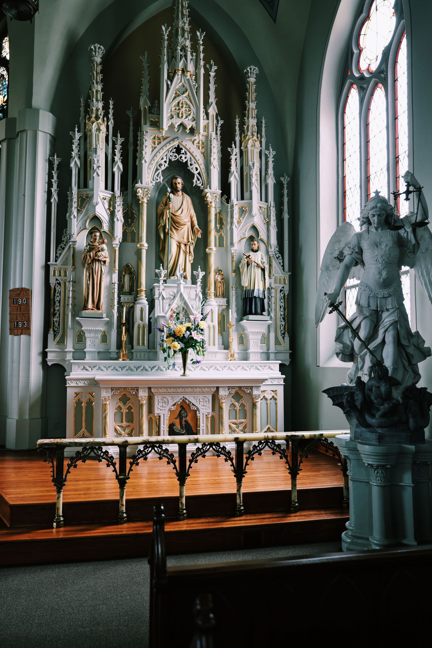 Ornate white Gothic church altar with religious statues, floral arrangement, and stained glass windows in a Catholic sanctuary.