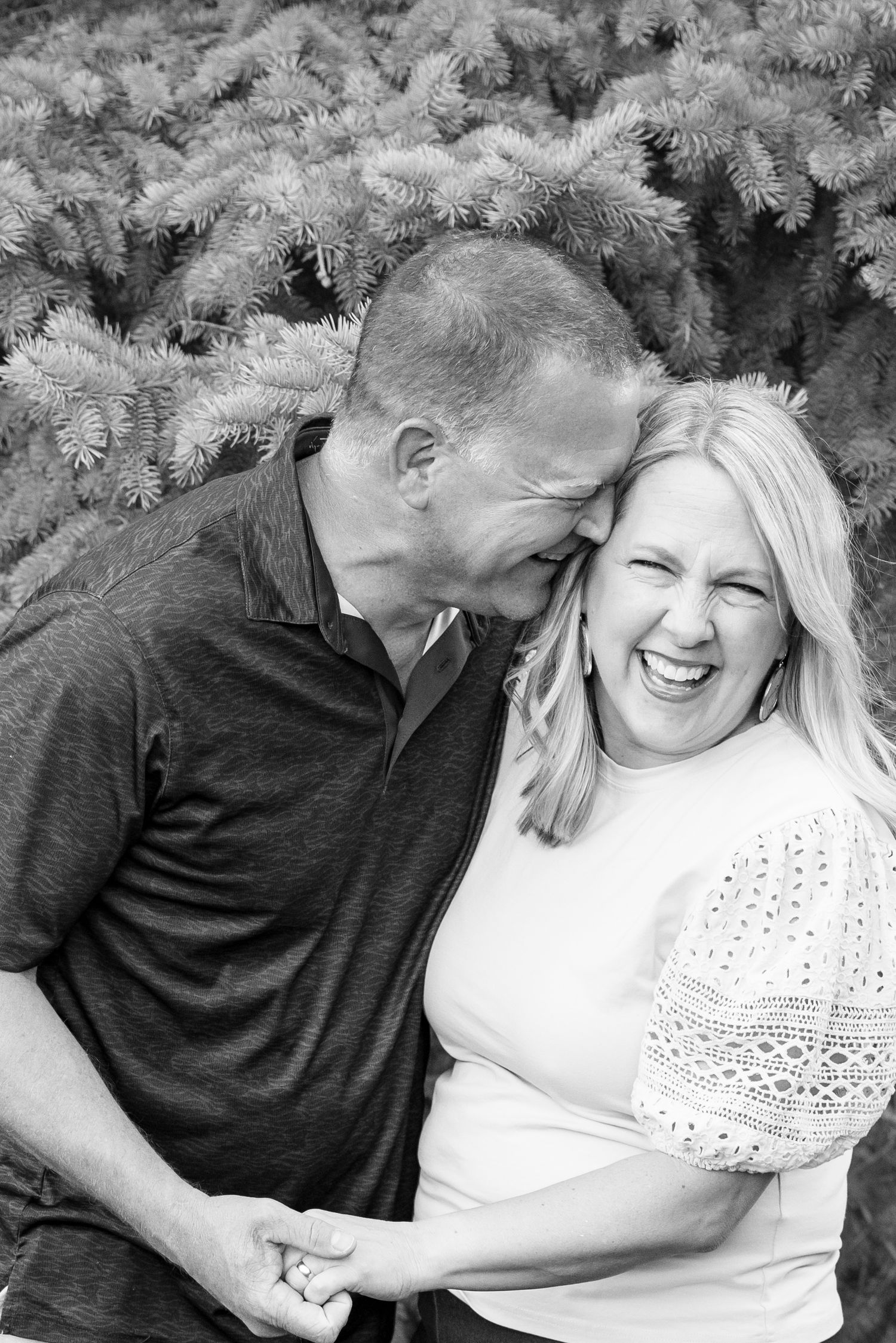 Black and white photo of a laughing couple holding hands in front of evergreen trees.