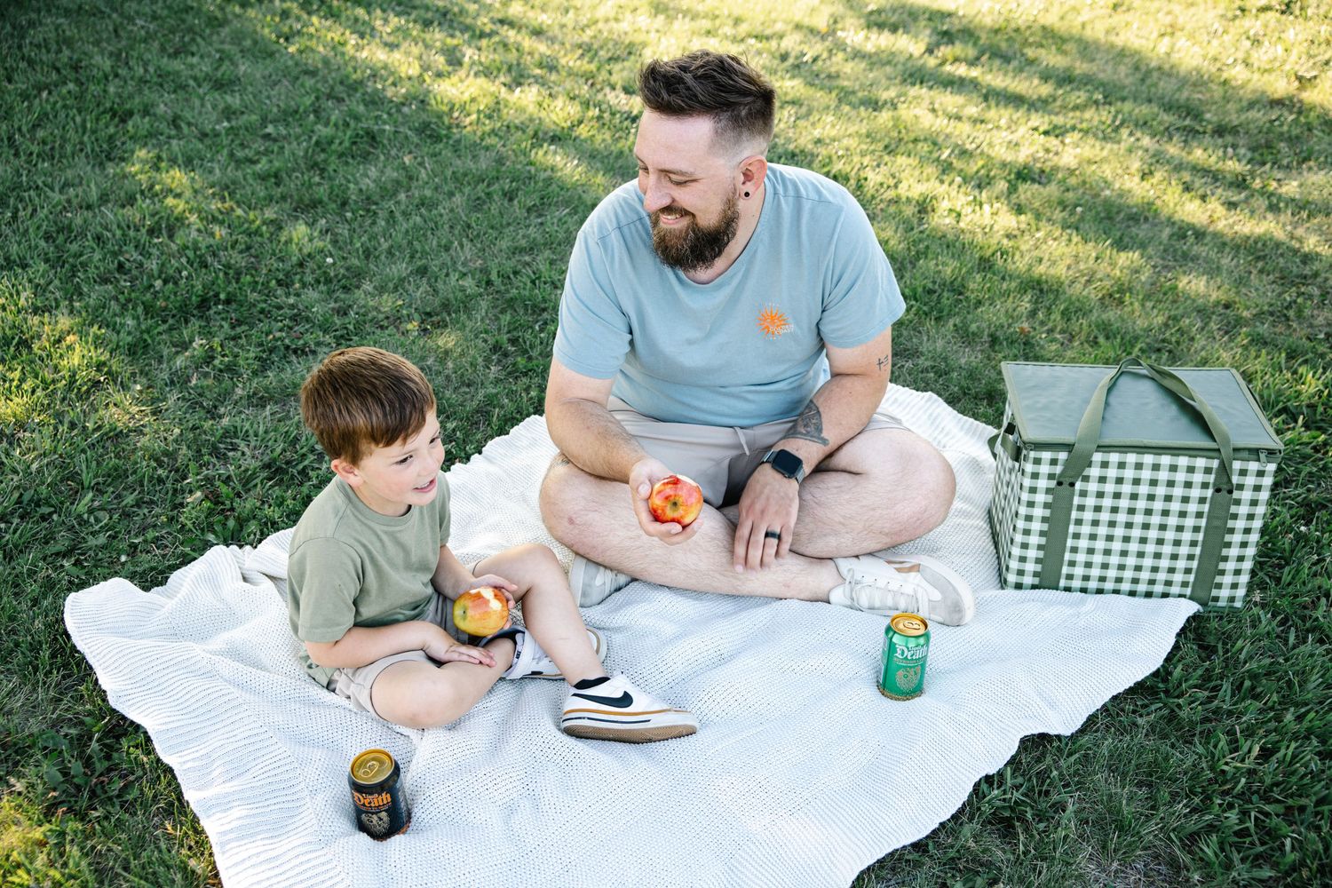 A father and young son enjoy a sunny outdoor picnic on a white blanket in a green grassy park.