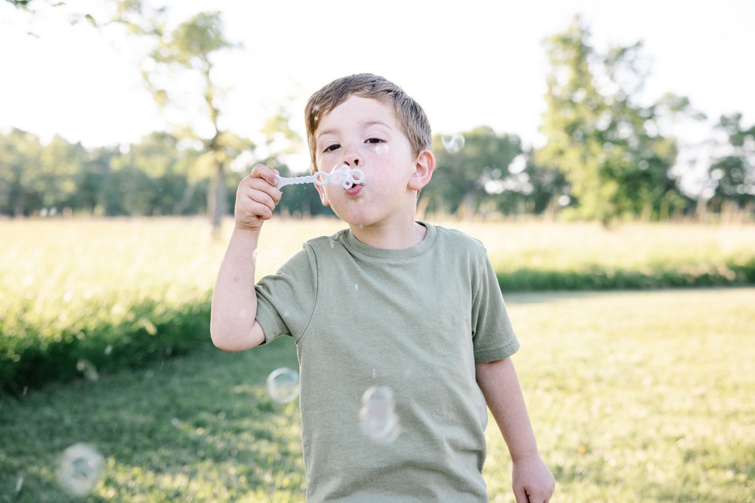 Young boy in green t-shirt blowing bubbles outdoors in a sunny park with lush green trees in background.