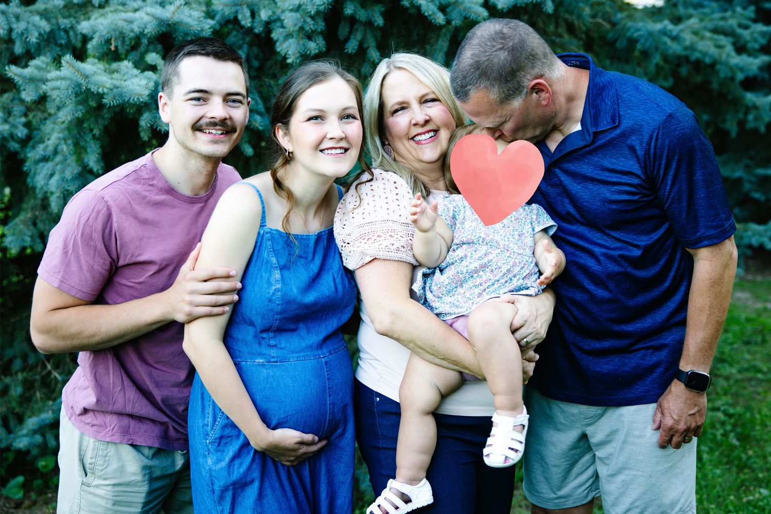 Happy family of four posing outdoors, pregnant woman in blue dress with partner and parents holding toddler with heart prop.