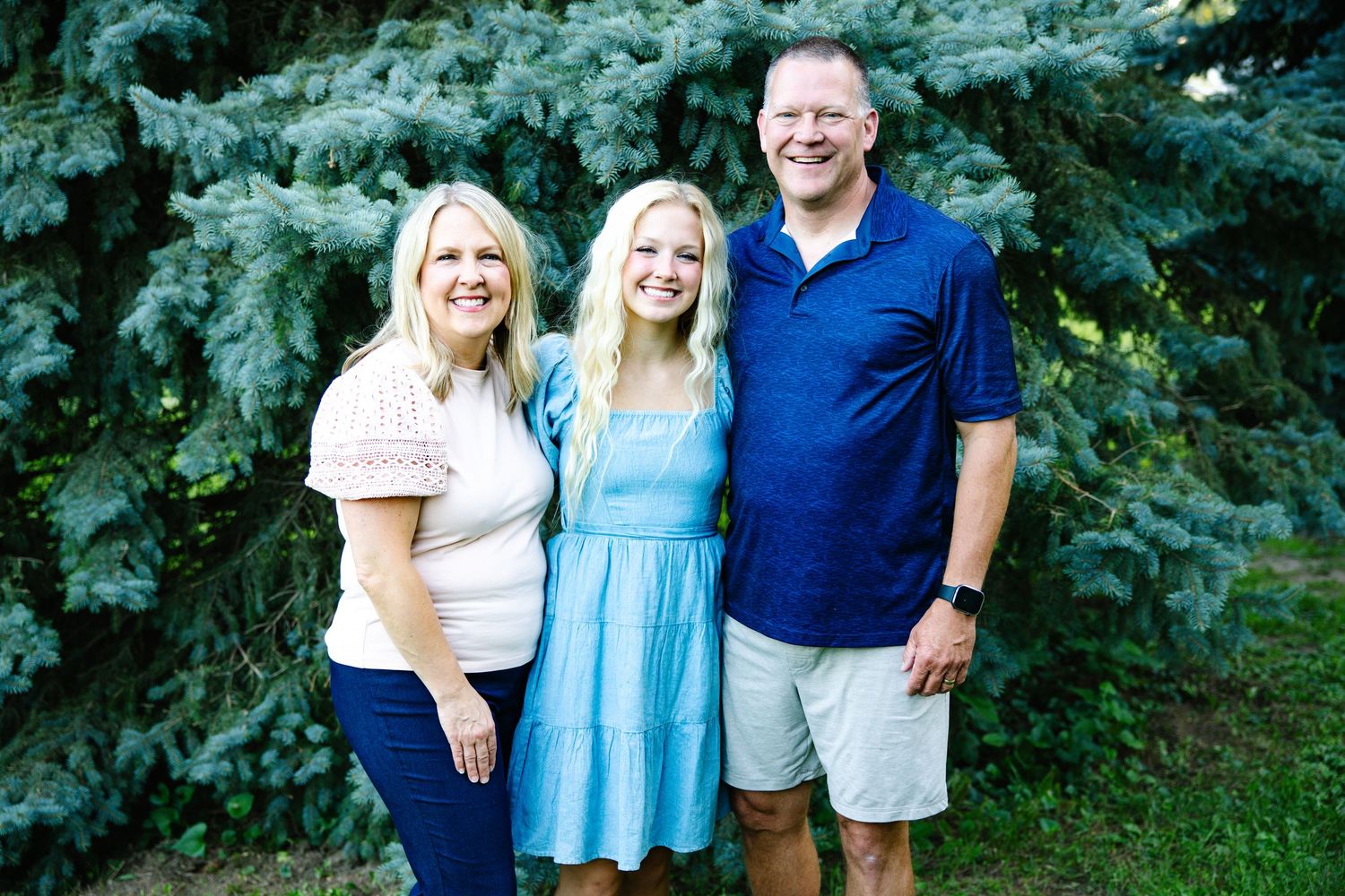 Happy family of three with teenage daughter smiling together outdoors near evergreen trees.