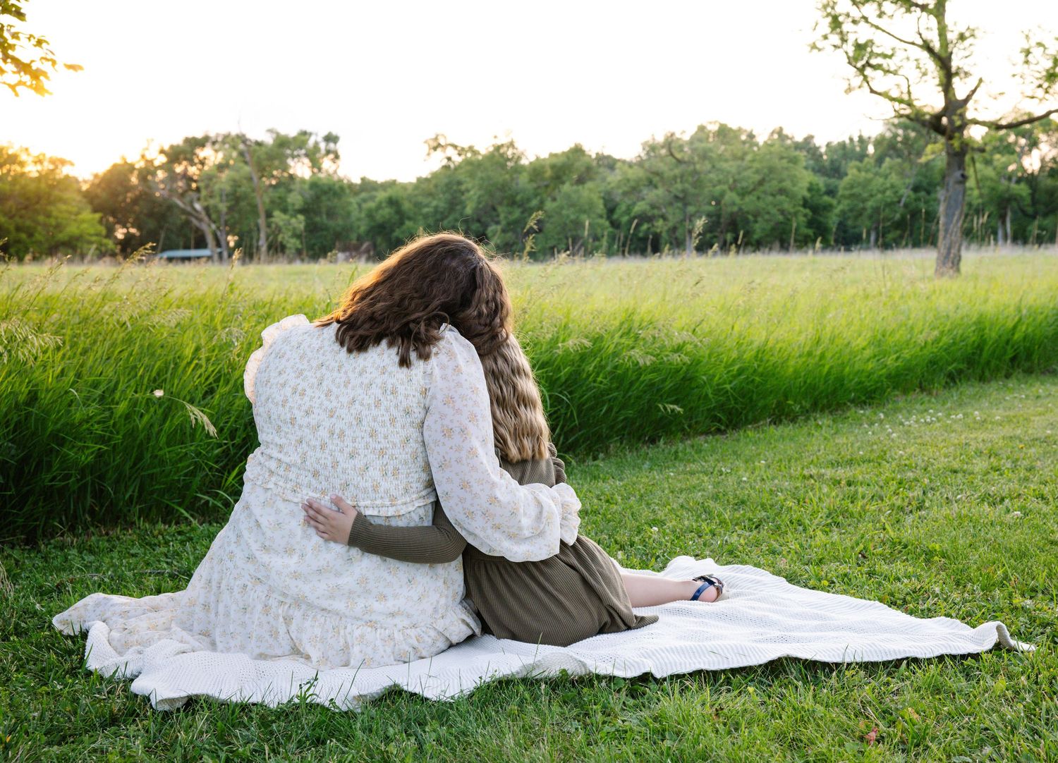 Two women sitting on a blanket in a green meadow at sunset, embracing each other while facing away from the camera.