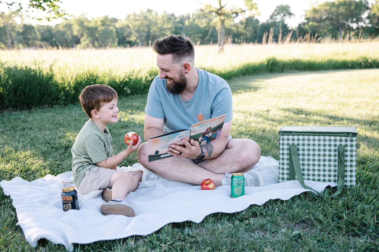 A bearded father reads a book with his young son during a relaxing outdoor picnic on a sunny day.