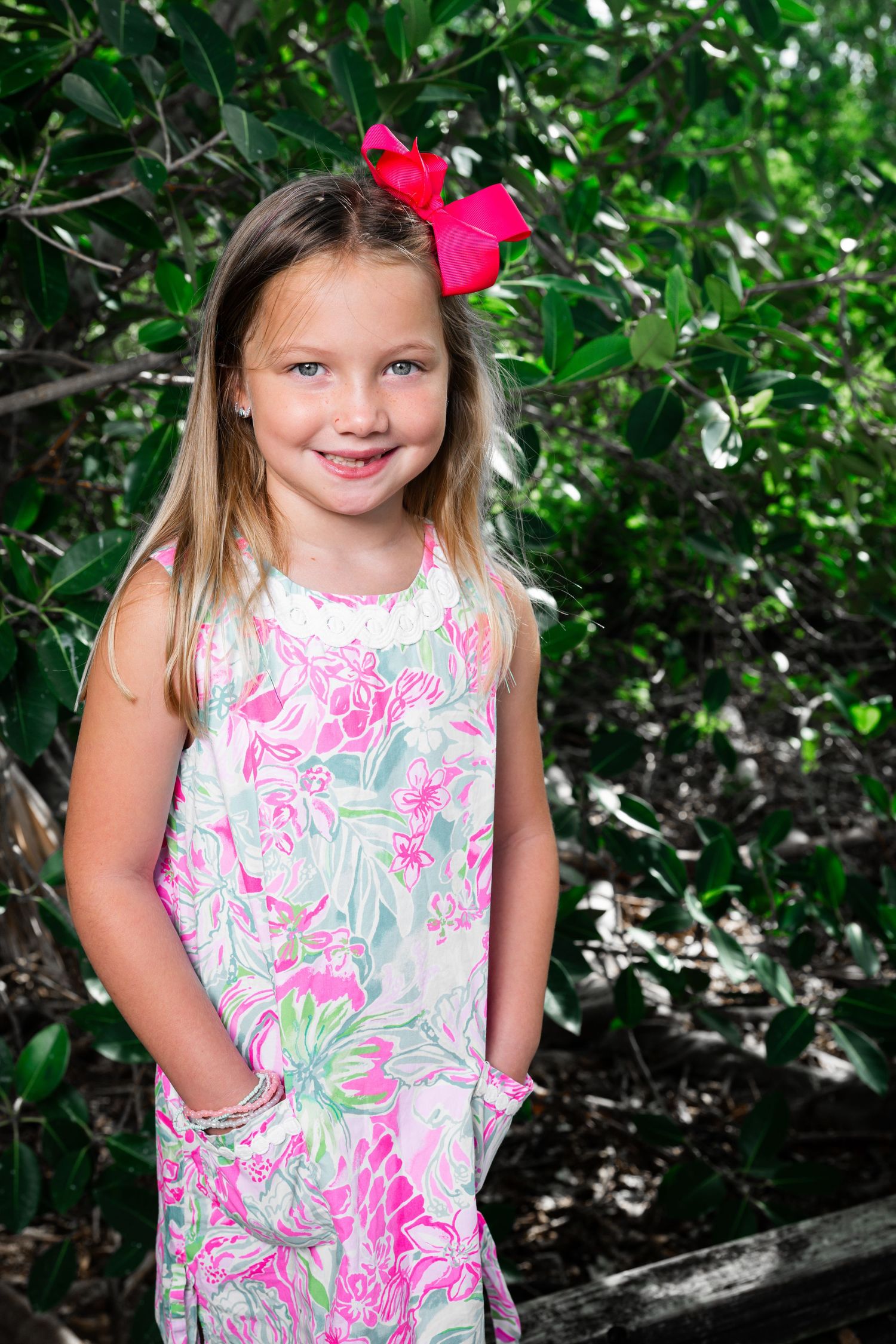 Young girl in floral dress with pink bow smiling outdoors among green foliage.