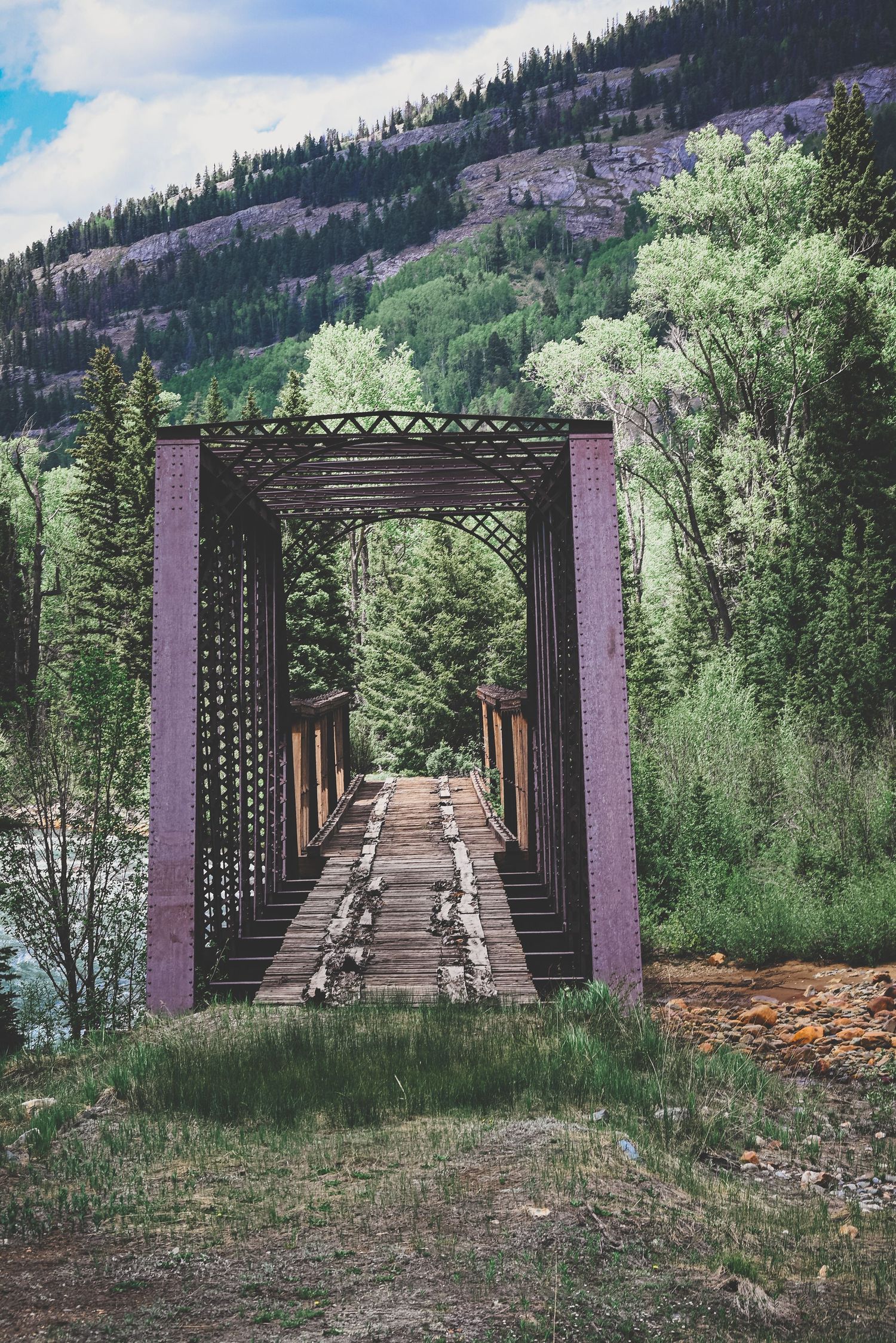 Rustic wooden pedestrian bridge with metal framework leading into a lush green mountain forest.