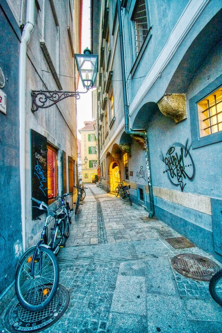Narrow cobblestone alley with bicycles, graffiti walls, and warm glowing lights in a European city.