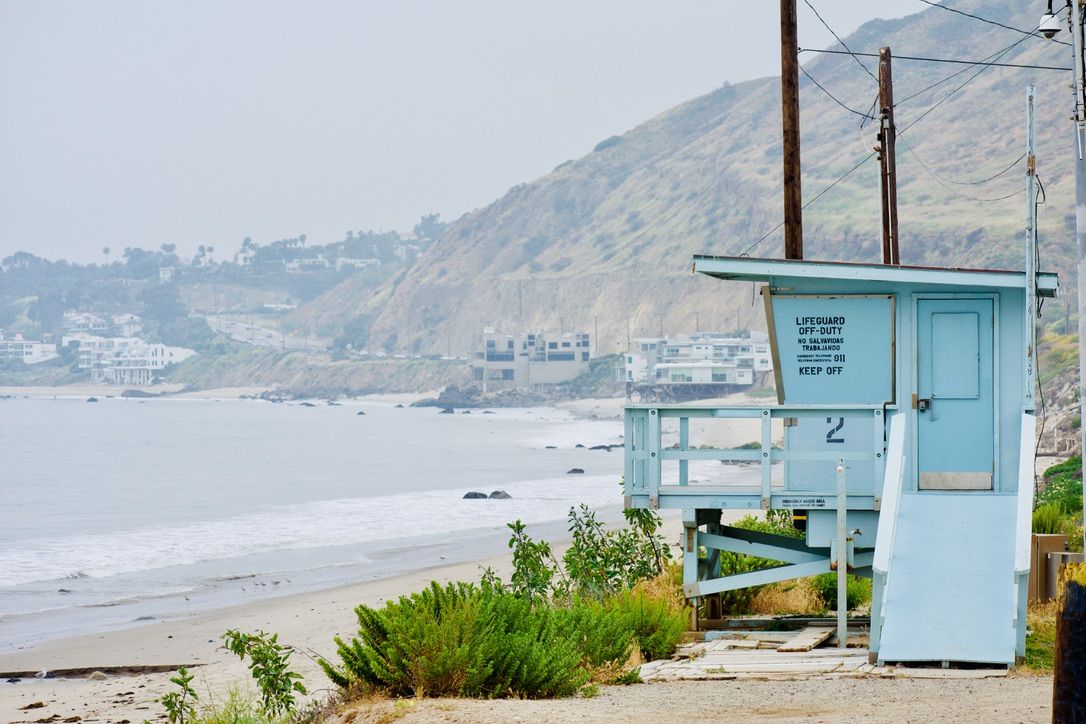 Light blue lifeguard station on a foggy Malibu beach with coastal cliffs in the background.