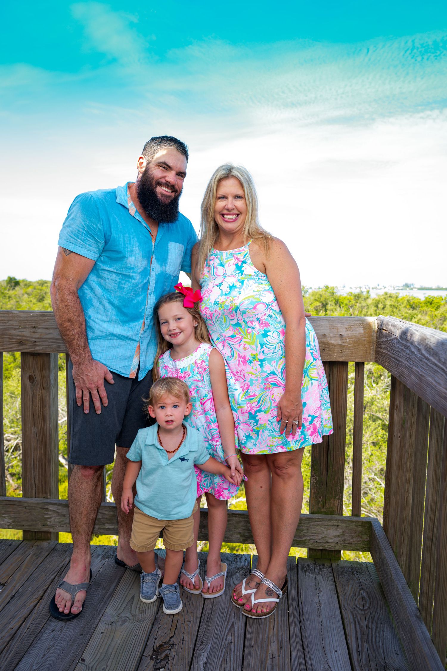 Family of four posing on wooden deck overlooking tropical greenery and blue sky at a beach destination.