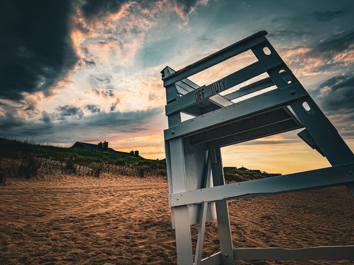 A white wooden lifeguard chair stands on sandy beach at dramatic sunset with colorful orange and dark stormy clouds overhead.