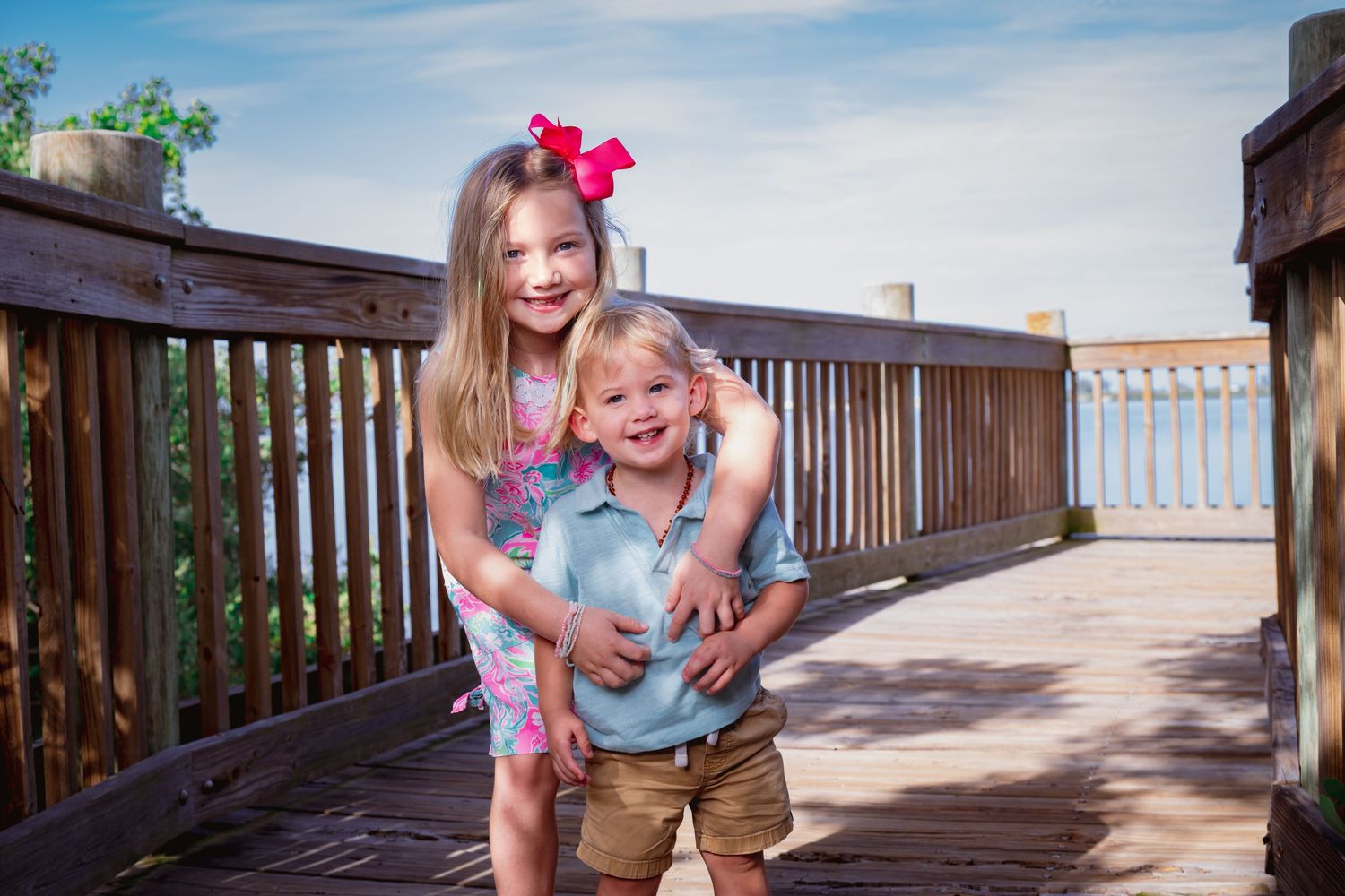 Two young siblings smiling and hugging on a wooden bridge outdoors on a sunny day.