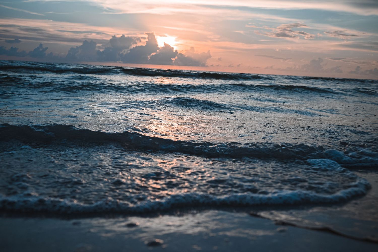 Soft ocean waves wash ashore on a peaceful beach during a warm pastel-colored sunset.