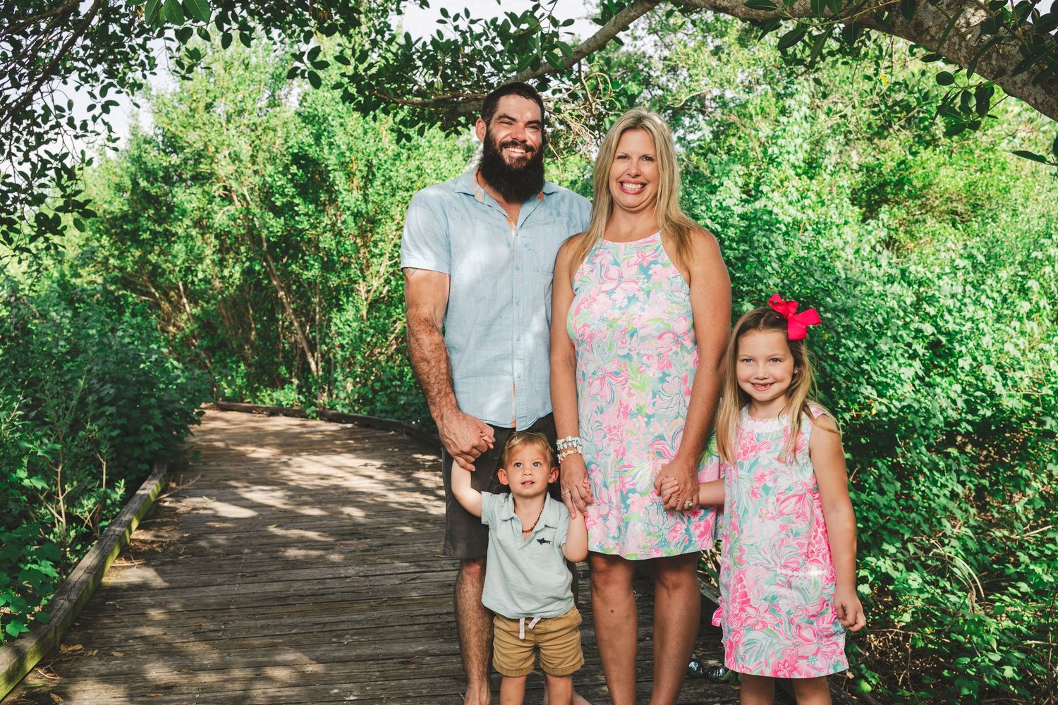Family of four poses on a wooden boardwalk surrounded by lush green trees and shrubs.