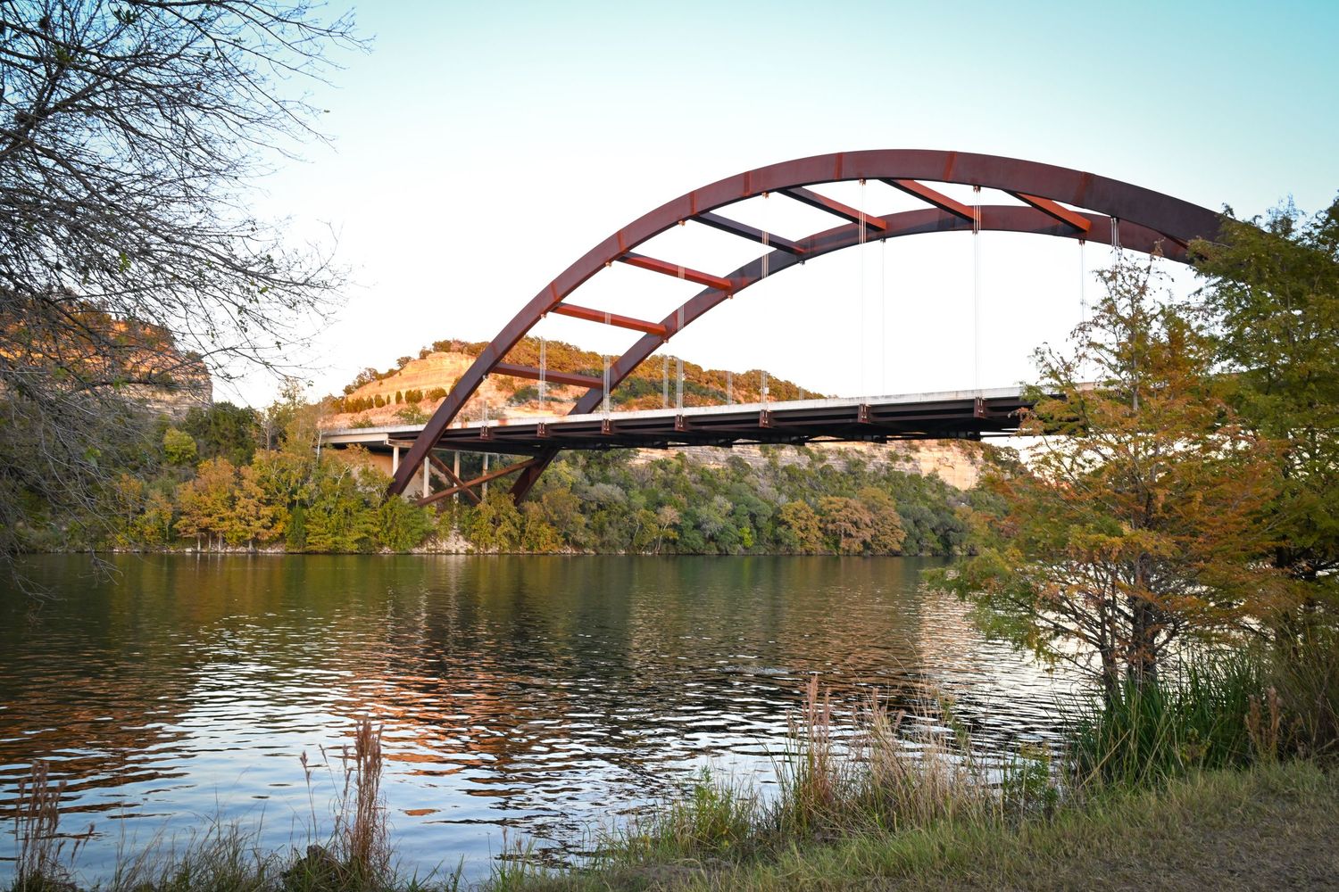The iconic Pennybacker 360 Bridge arching over calm river waters surrounded by autumn trees at dusk.