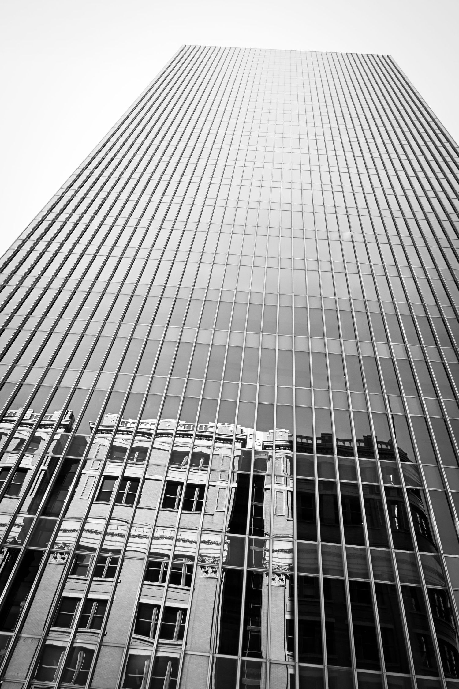 Black and white low-angle view of a tall glass skyscraper reflecting an older building below.
