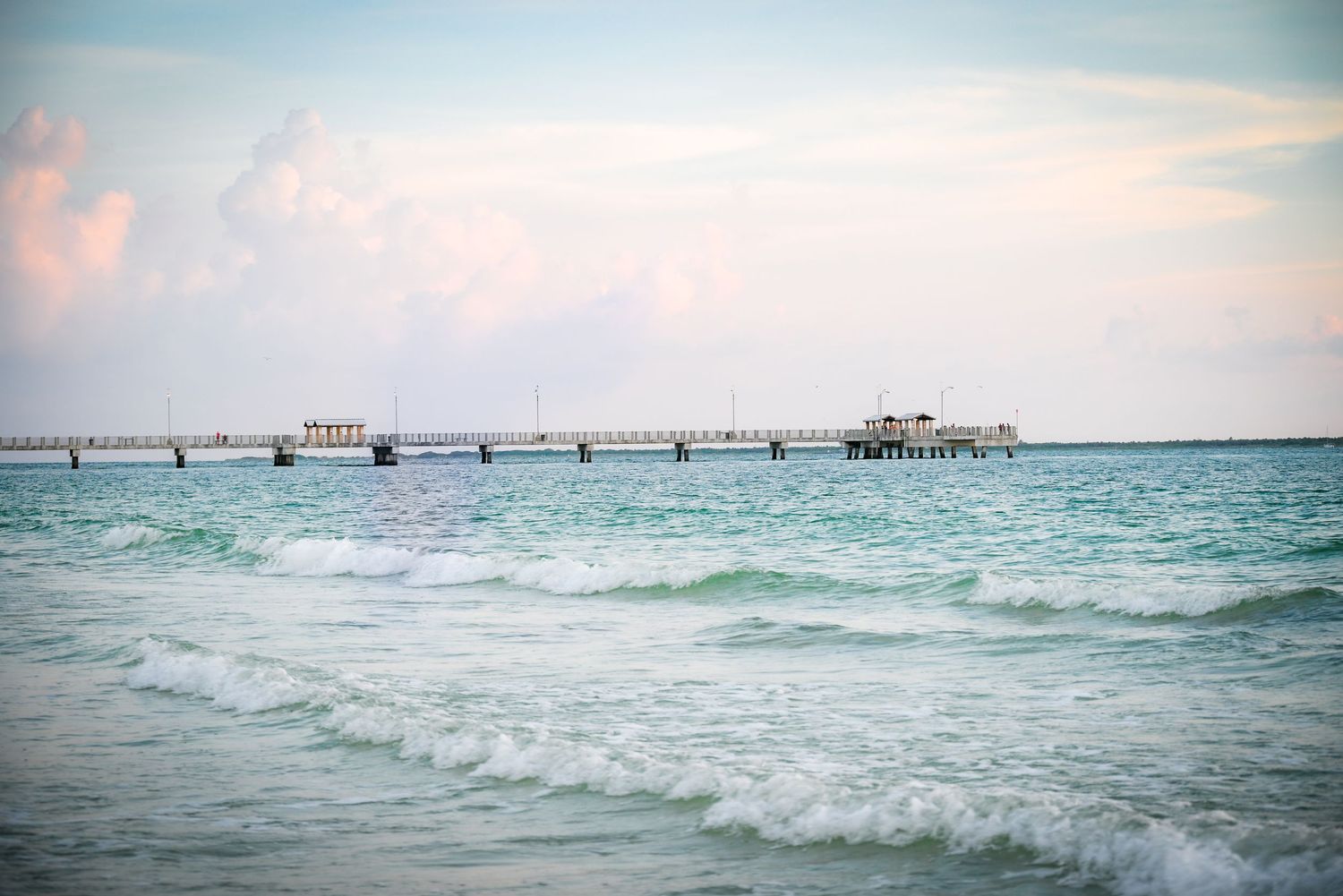 Turquoise ocean waves roll toward shore with a long fishing pier stretching into the sea.